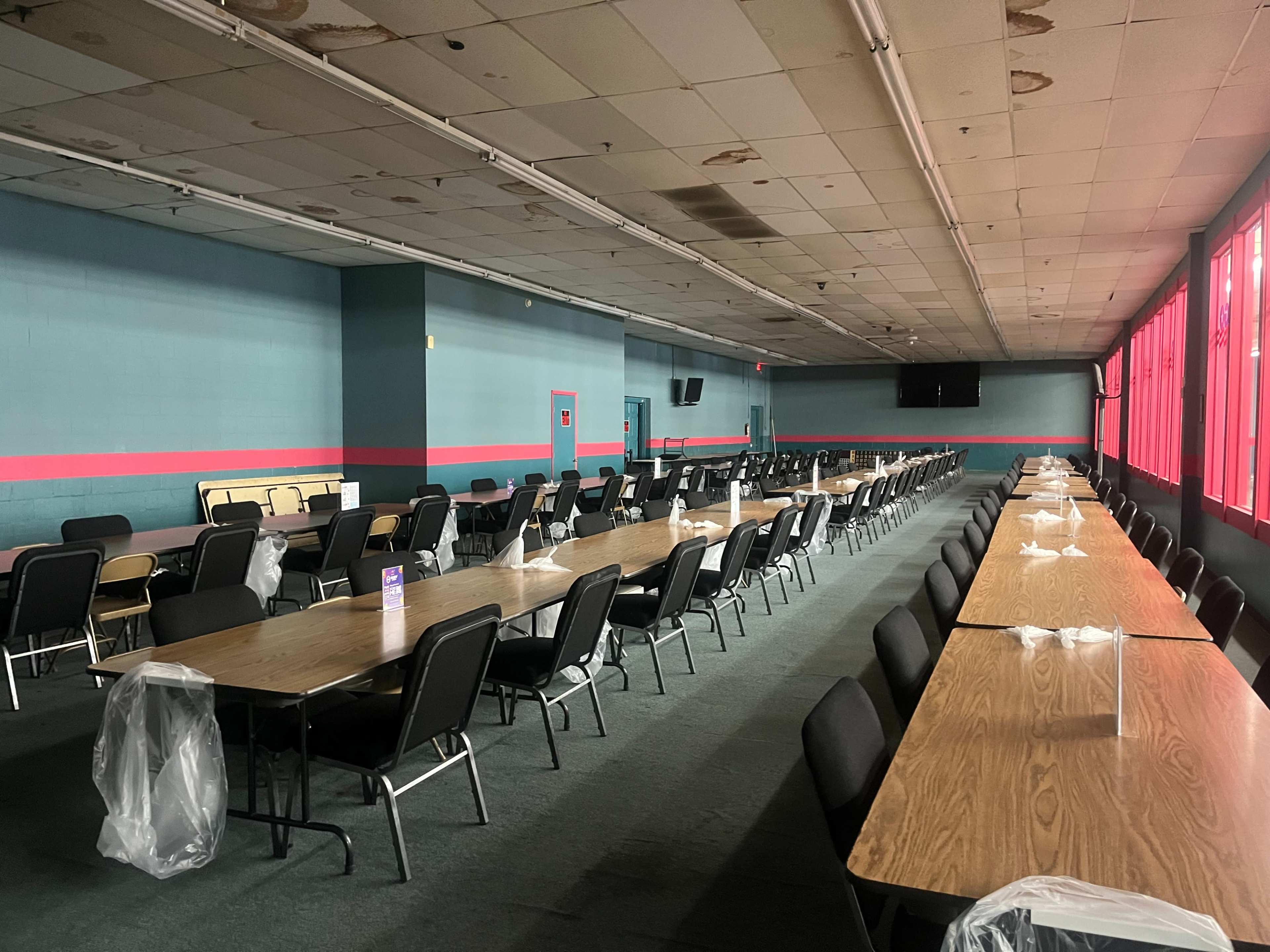 A long, empty banquet hall with rows of wooden tables and chairs, and large windows covered by red drapes.