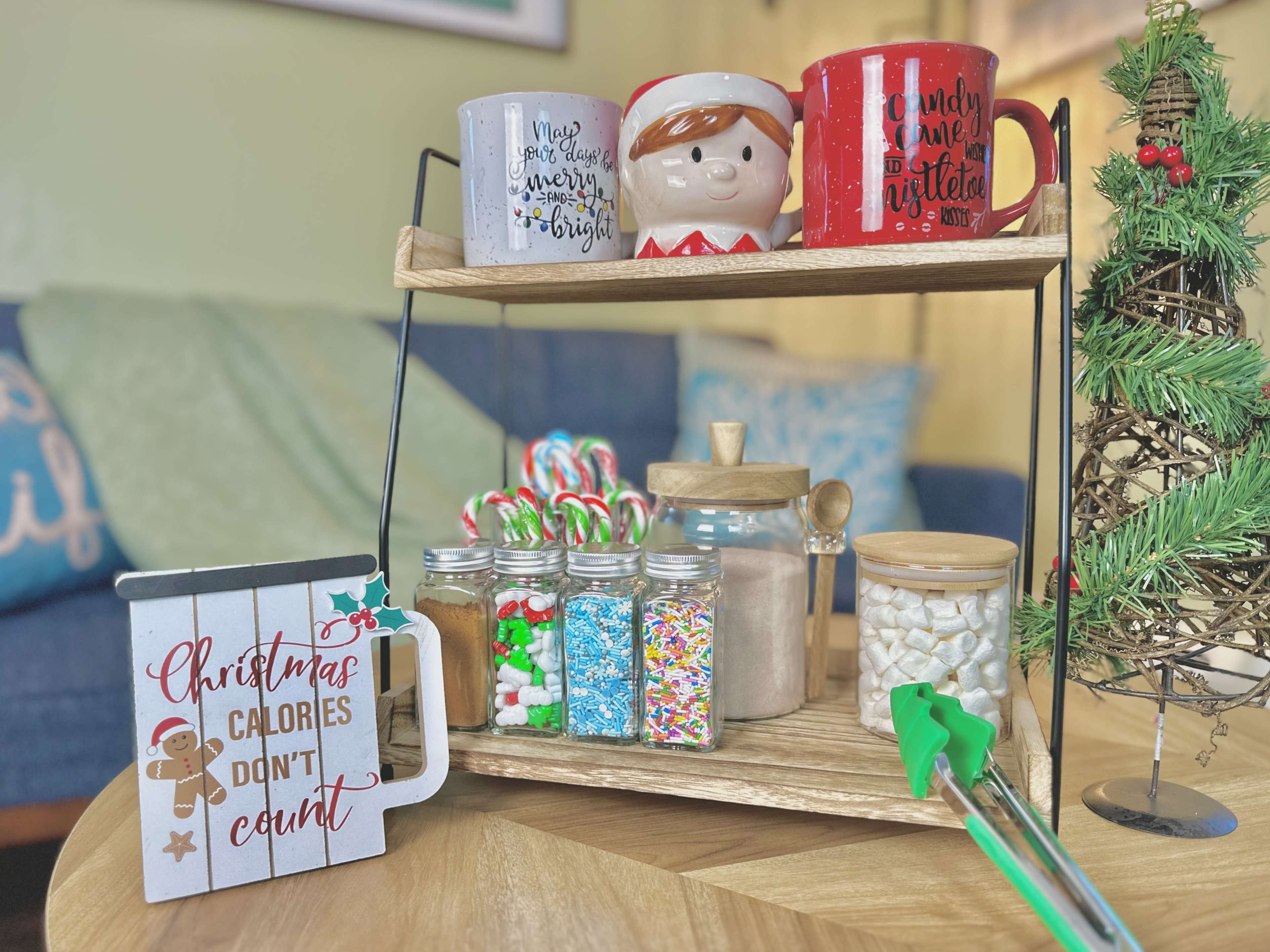 The image shows a festive display of holiday-themed mugs, candy canes, and jars filled with colorful baking supplies on a wooden shelf, accompanied by a decorative sign and a small Christmas tree.