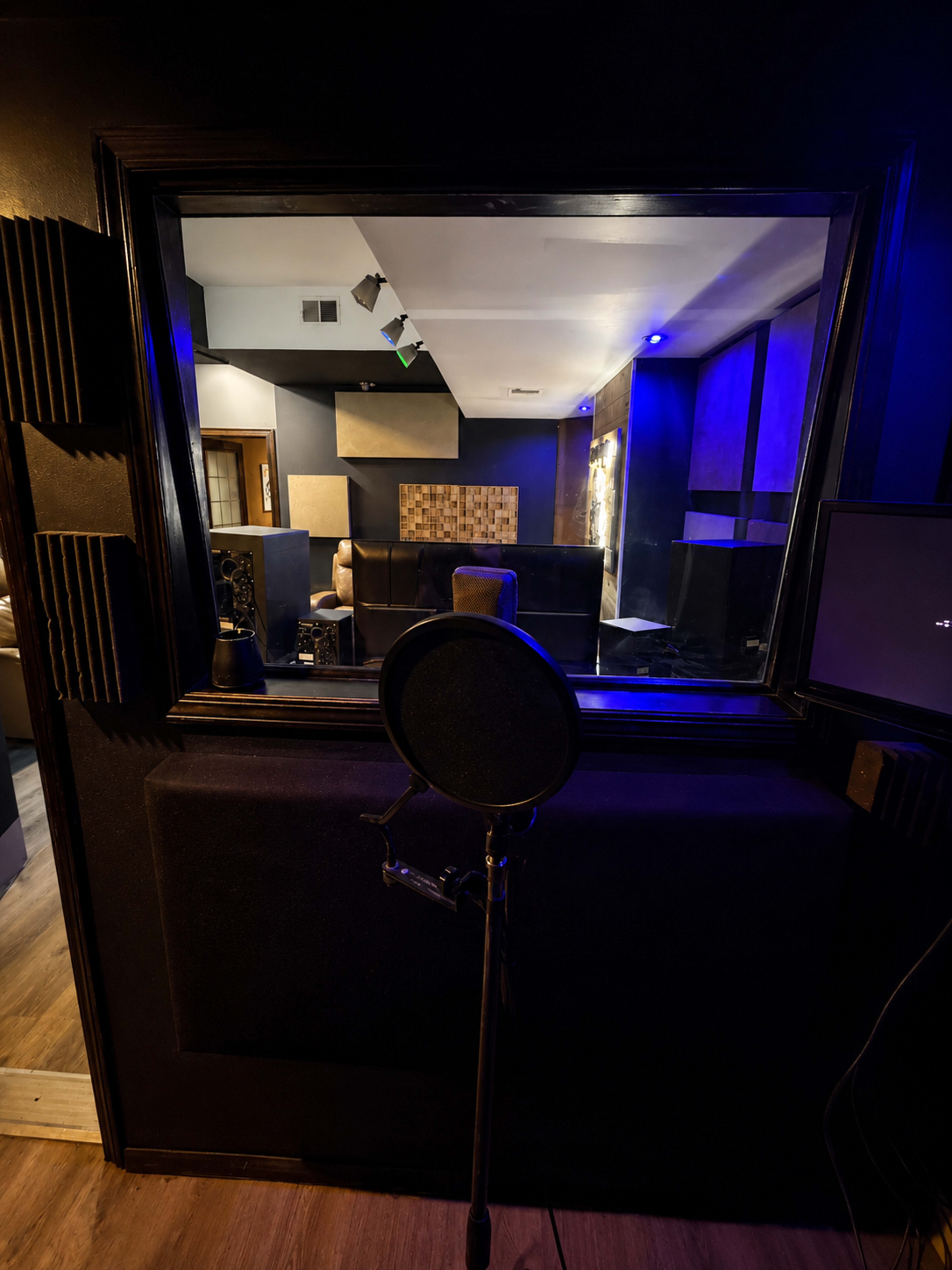 The image shows a recording studio with a microphone set up in front of a soundproof glass window, leading into a darkened room with soft lighting and a couch.