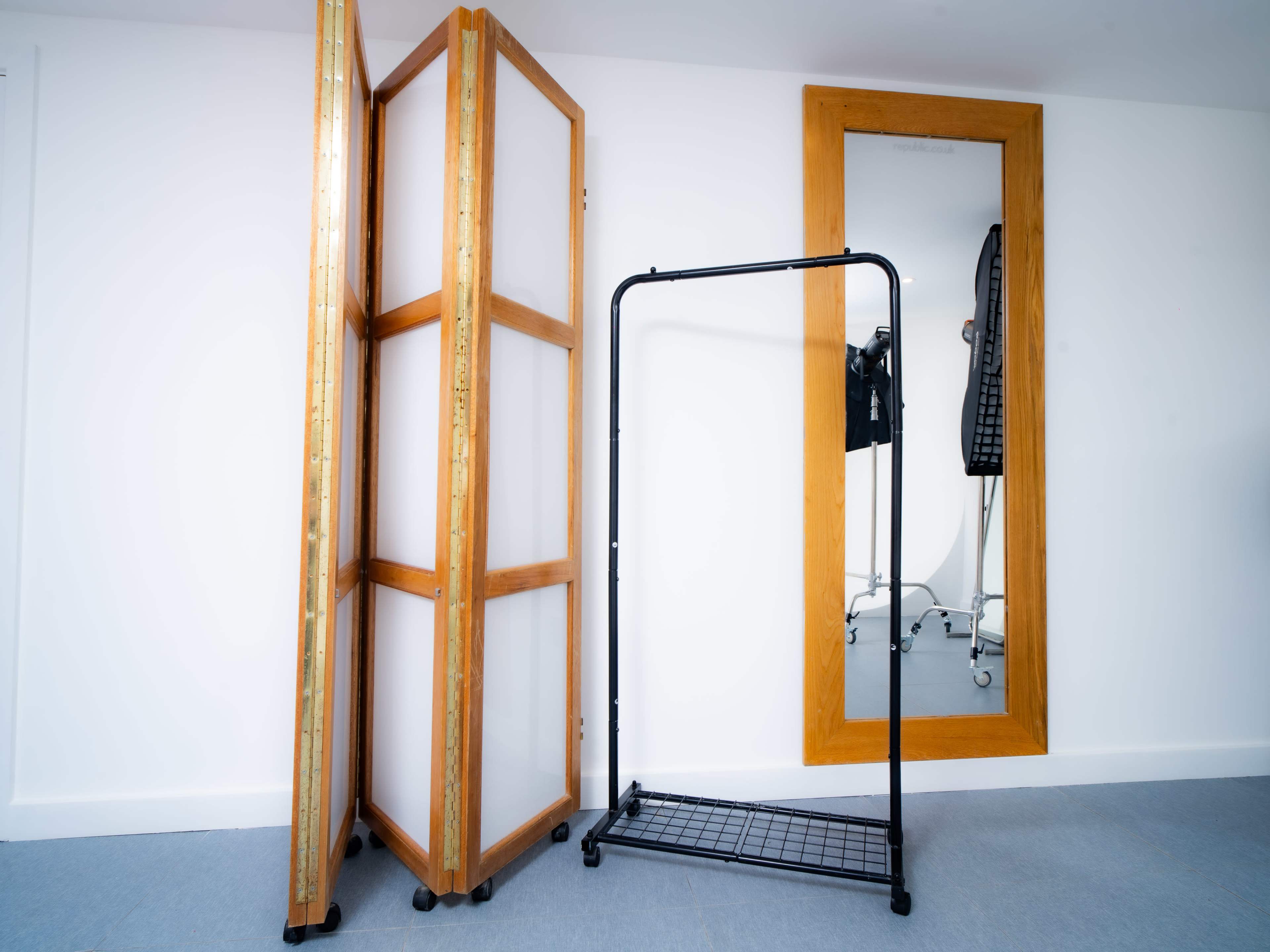 The image shows a room with a wooden folding screen, a black clothing rack, and a full-length mirror against a white wall.