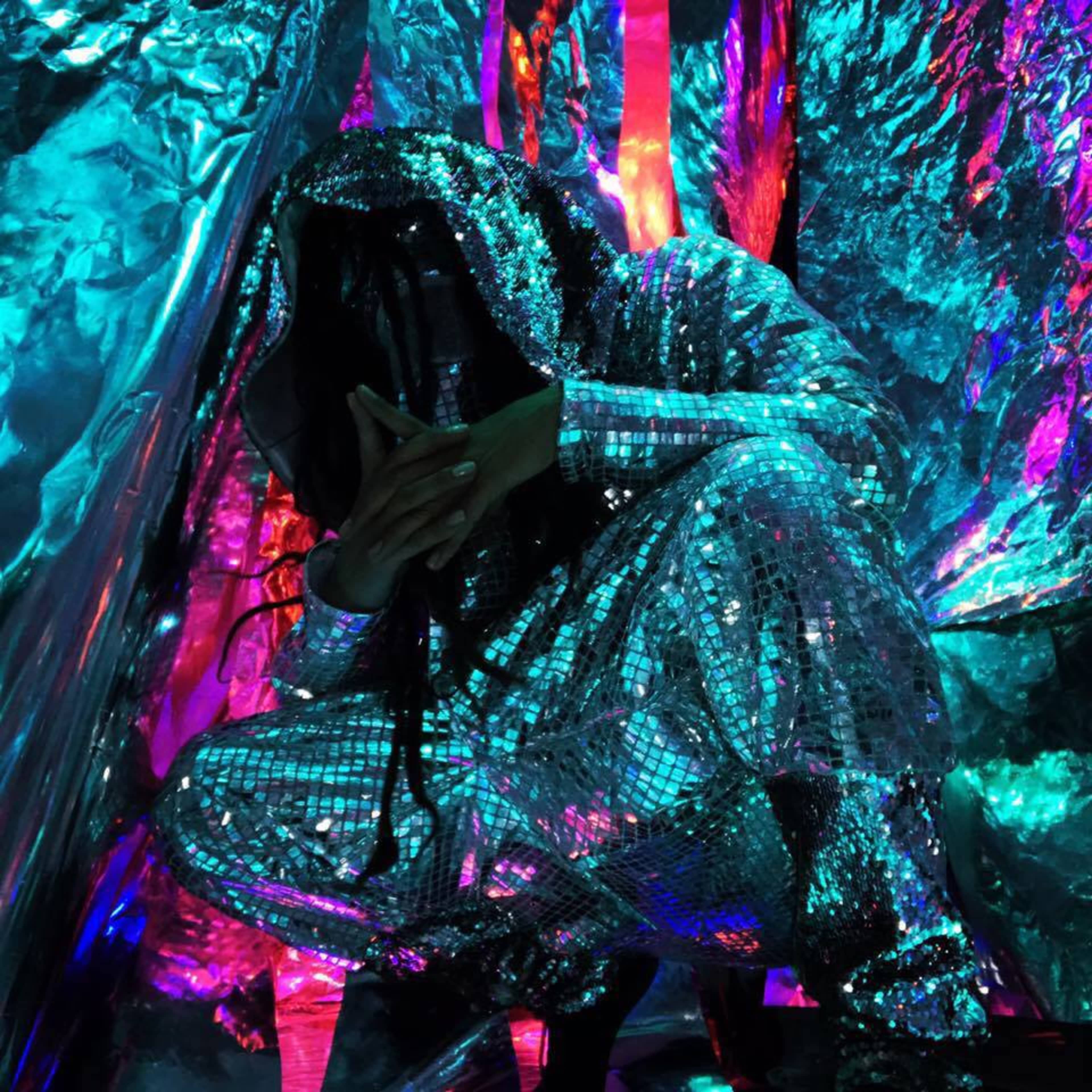 A person in a reflective, sequined outfit crouches among colorful, metallic backdrops.