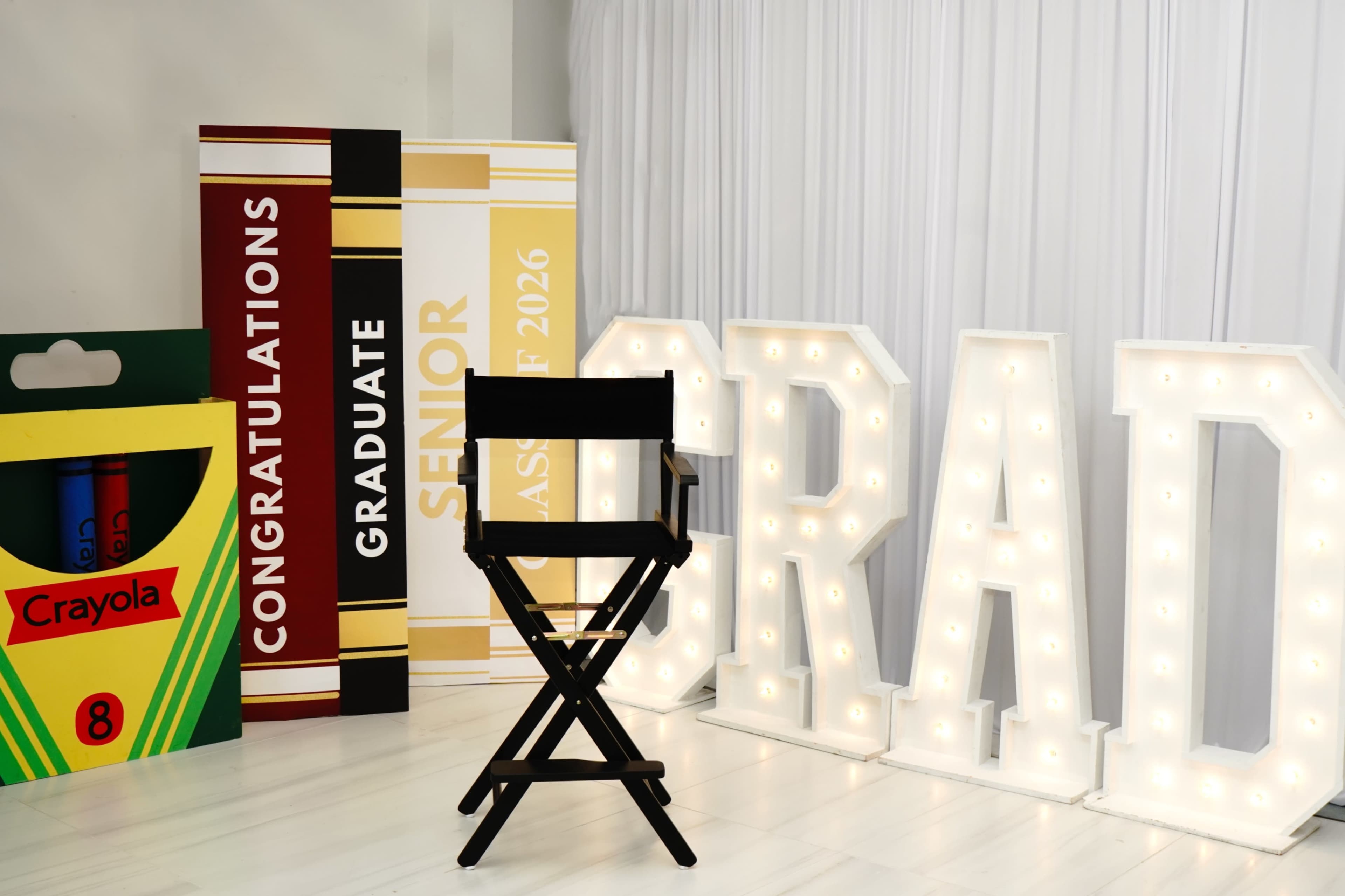 The image features a graduation-themed setup with illuminated letters spelling "GRAD," a black director's chair, and large signs that read "CONGRATULATIONS," "GRADUATE," and "SENIOR CLASS OF 2026," along with a Crayola box of crayons.