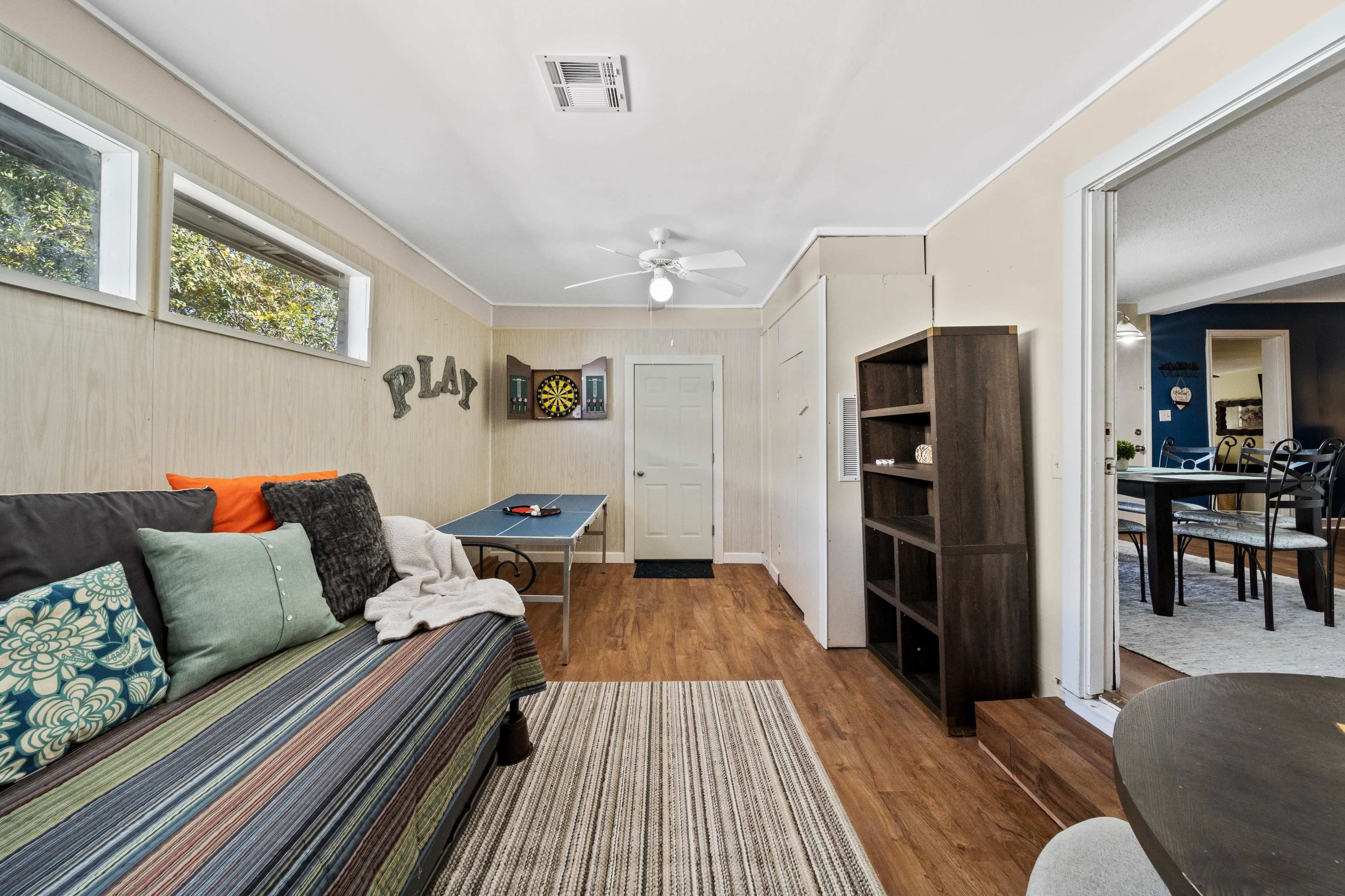 The image shows a brightly lit indoor space featuring a couch, a dartboard, a table, and an open doorway leading to another room.