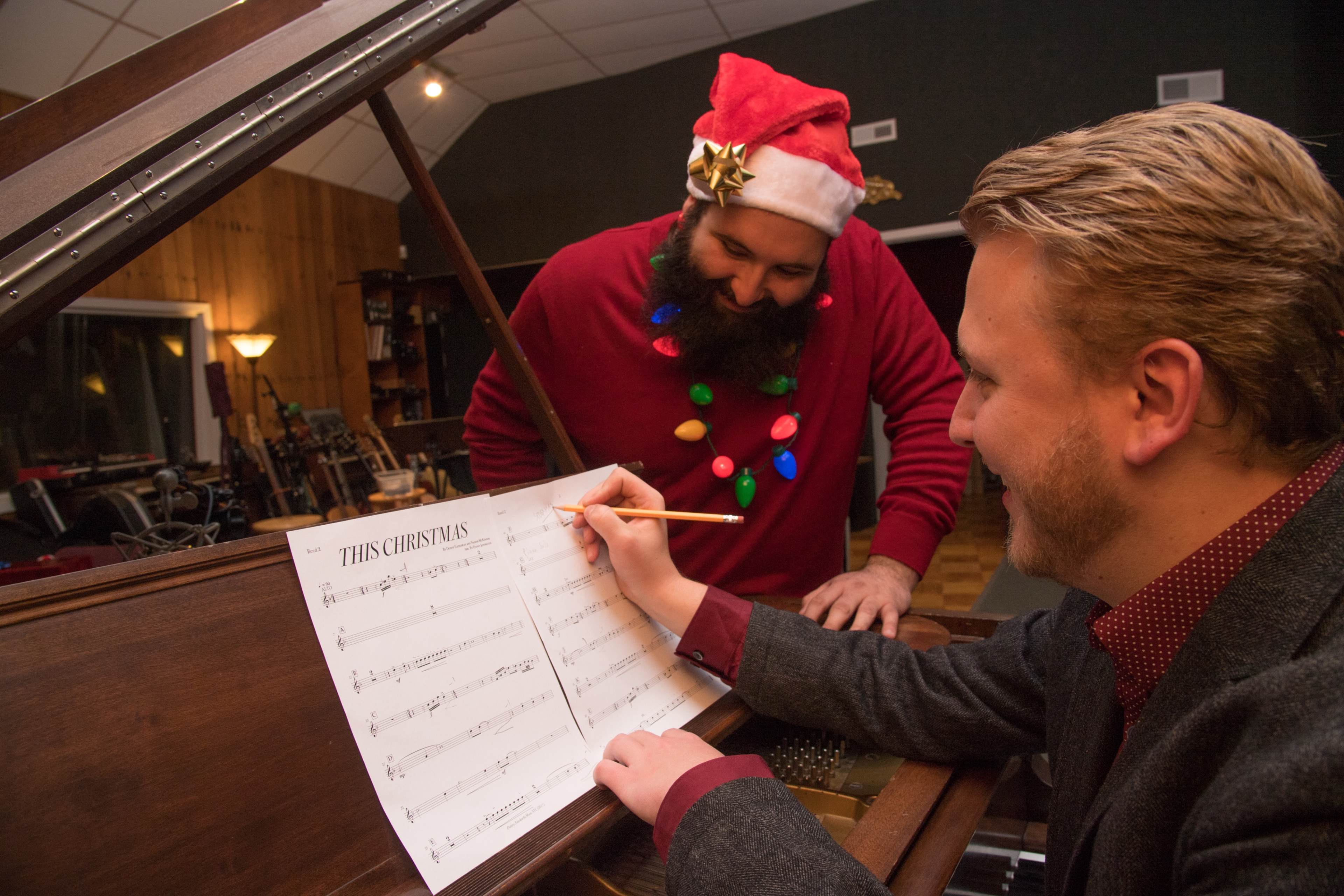 Two men are in a recording studio, with one wearing a Santa hat and festive sweater, as they look over sheet music on a grand piano.