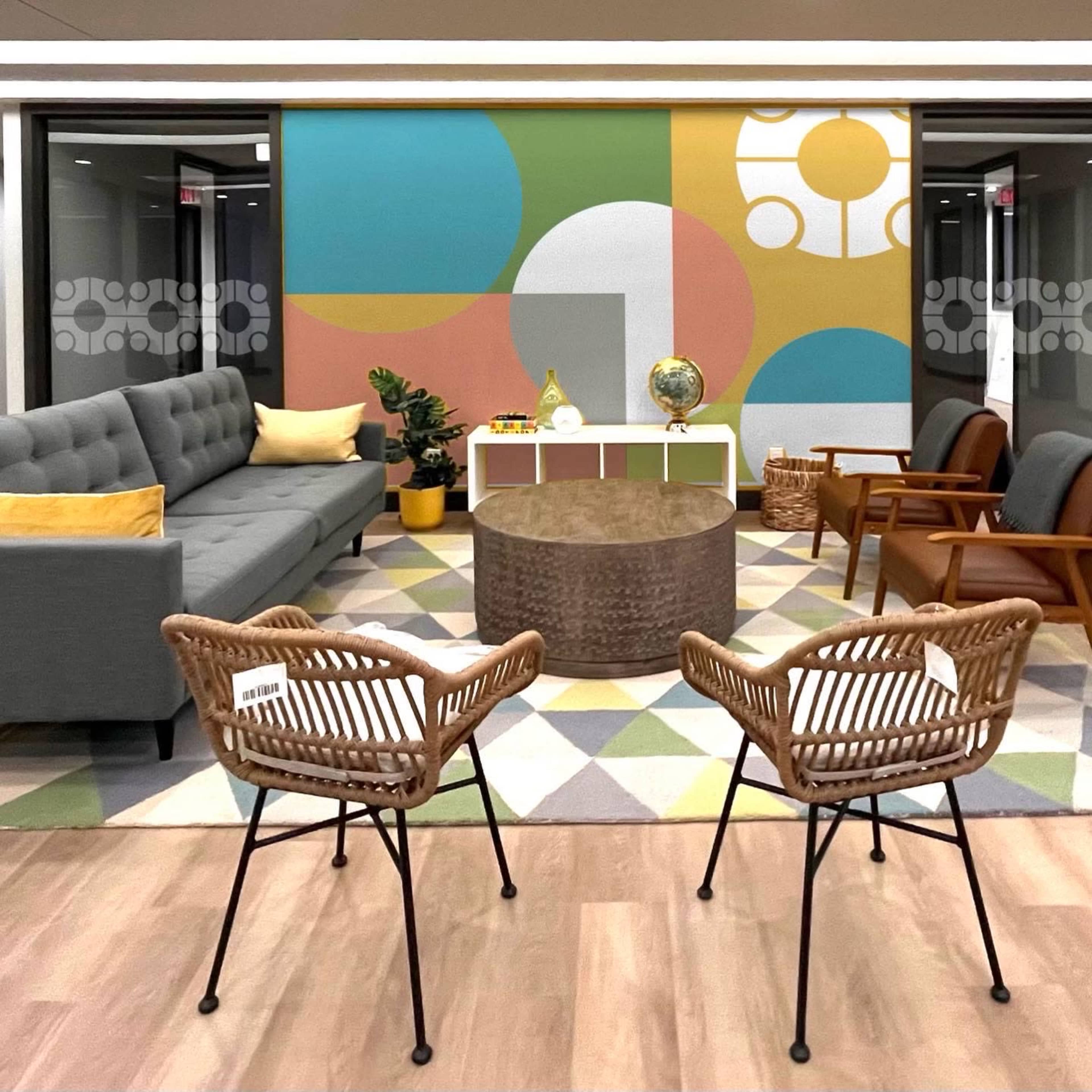 A modern lounge area featuring a gray sofa, two wicker chairs, a round coffee table, and a colorful geometric wall mural.