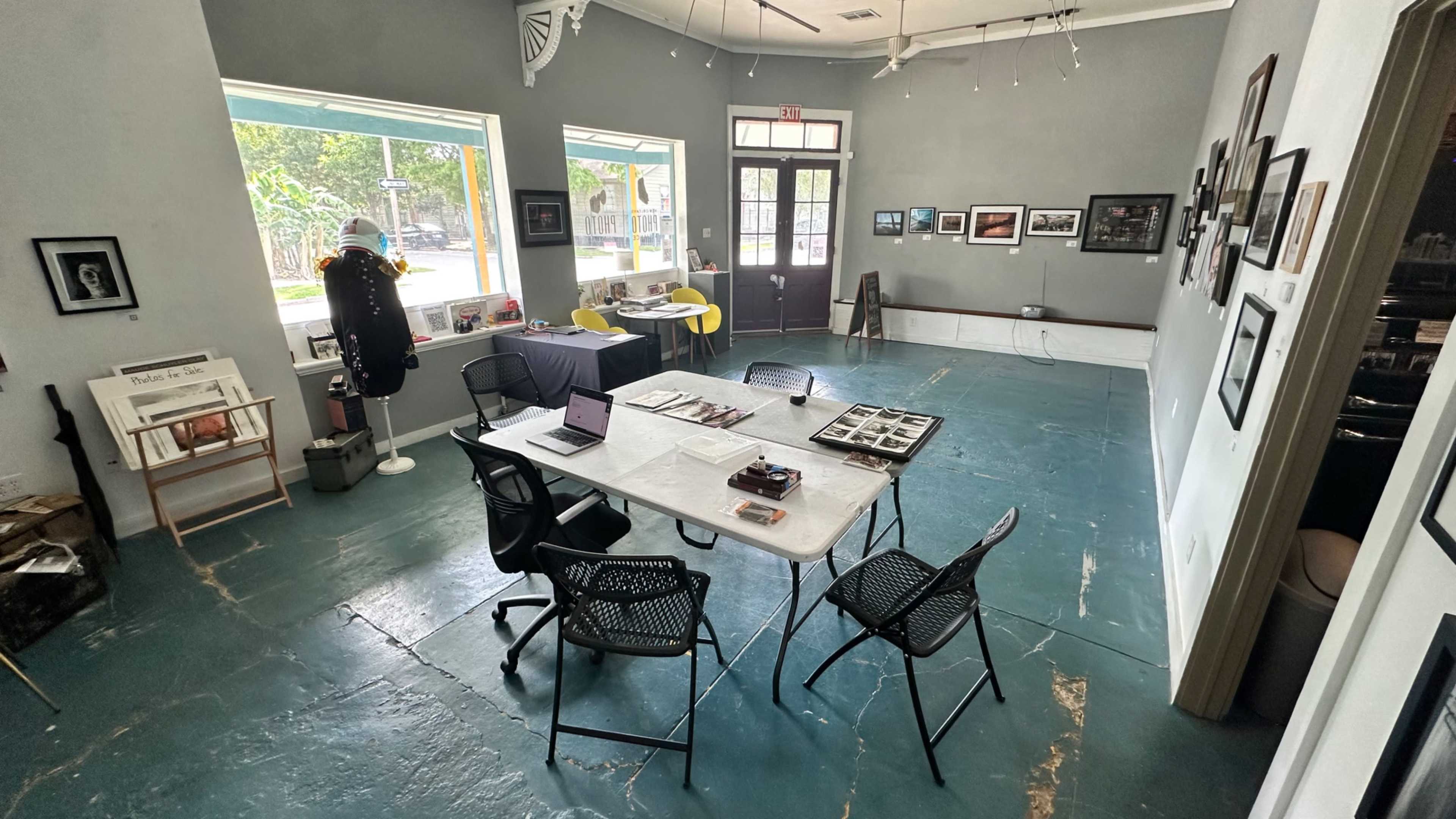 The image shows an art gallery with a table and chairs set up in the center, displaying photography and other art pieces on the walls.