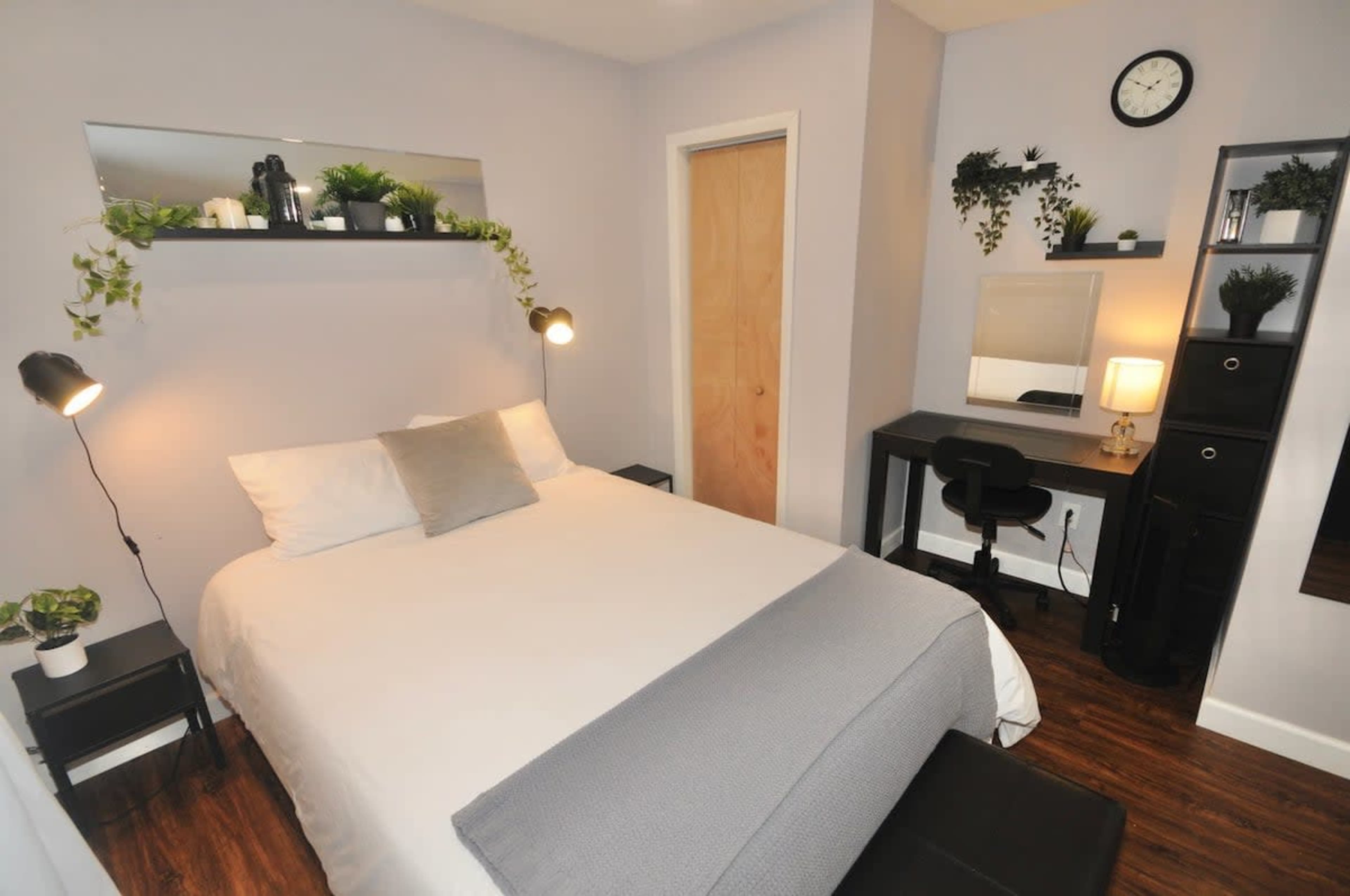 A modern bedroom with a double bed, a desk with a chair, and storage units, all accented by wall shelves with plants.