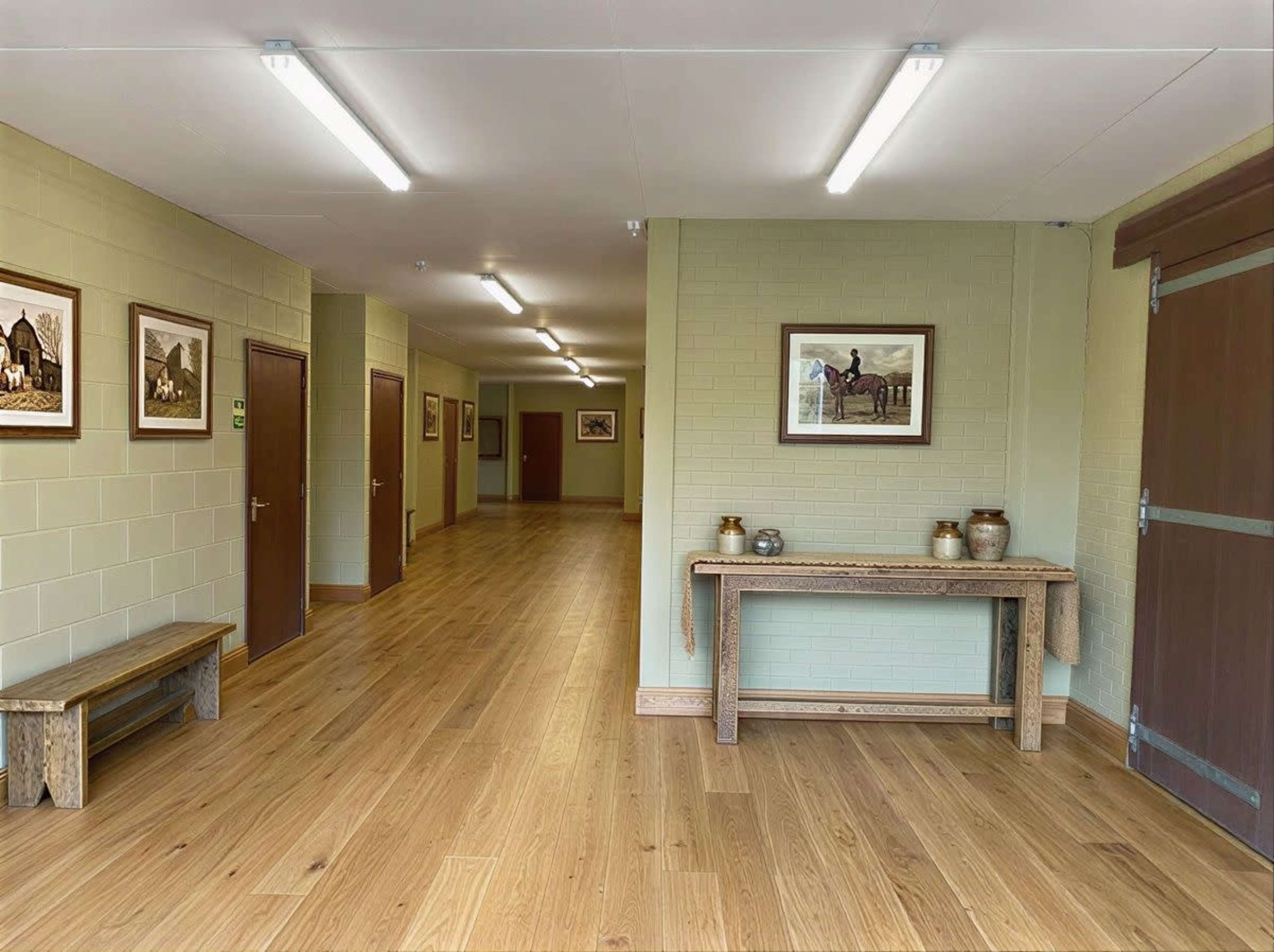 The image shows a well-lit hallway with wooden flooring, featuring framed photographs on the walls and a bench along one side.