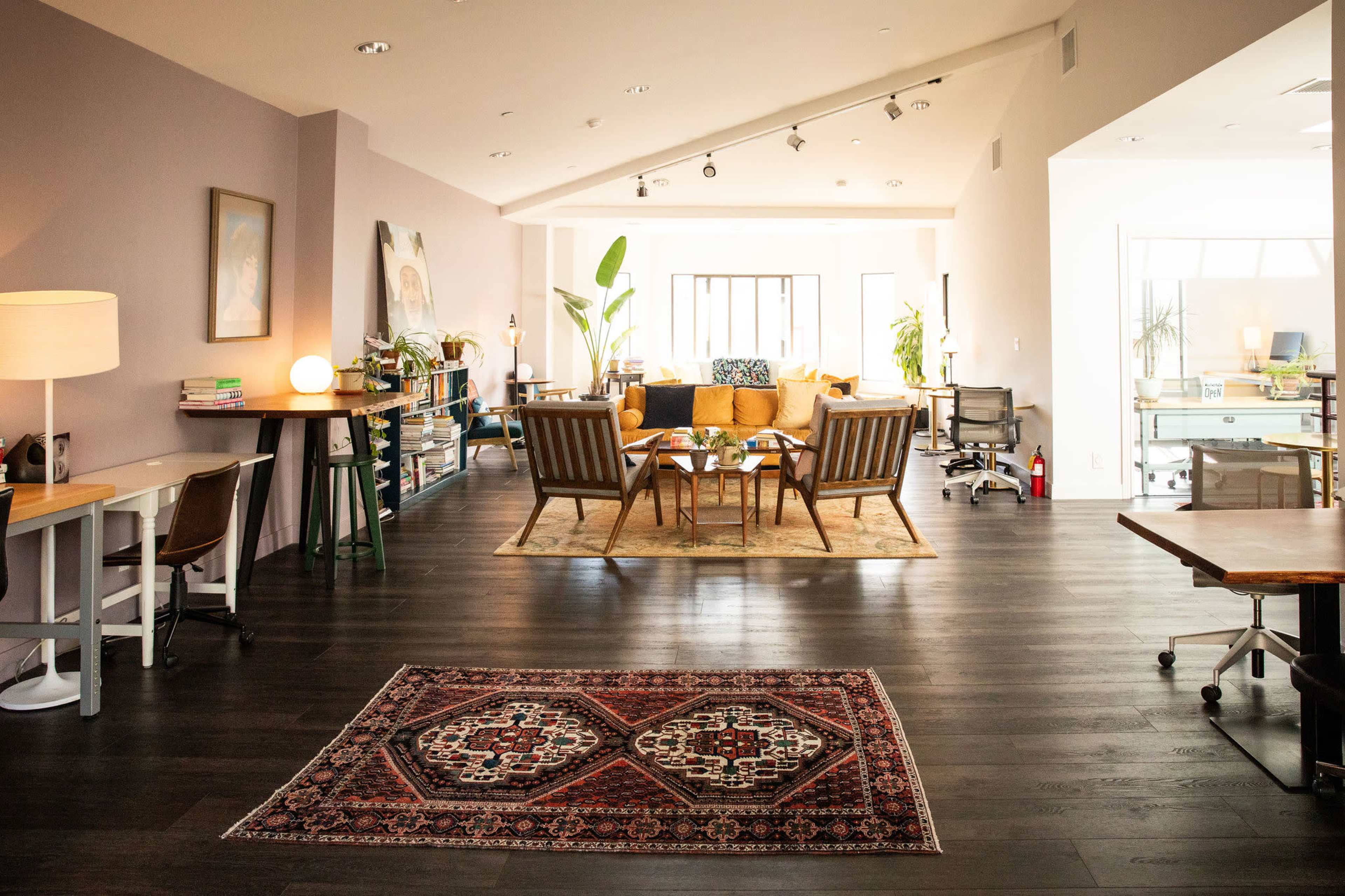 The image depicts a spacious, modern office space featuring a lounge area with a large rug, comfortable seating, and workstations along the sides.