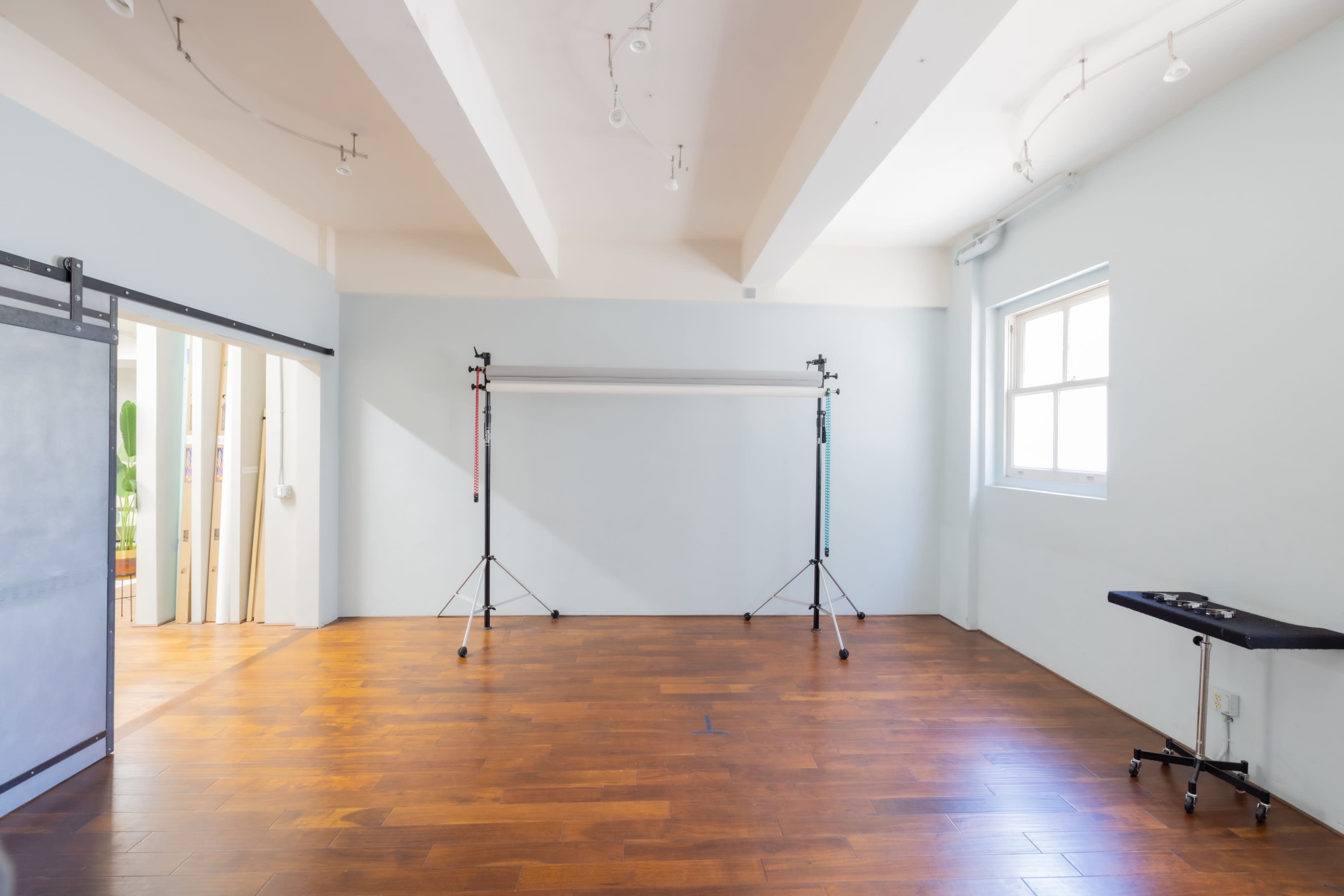 The image shows a spacious, empty room with wooden floors, white walls, and a few studio equipment stands.
