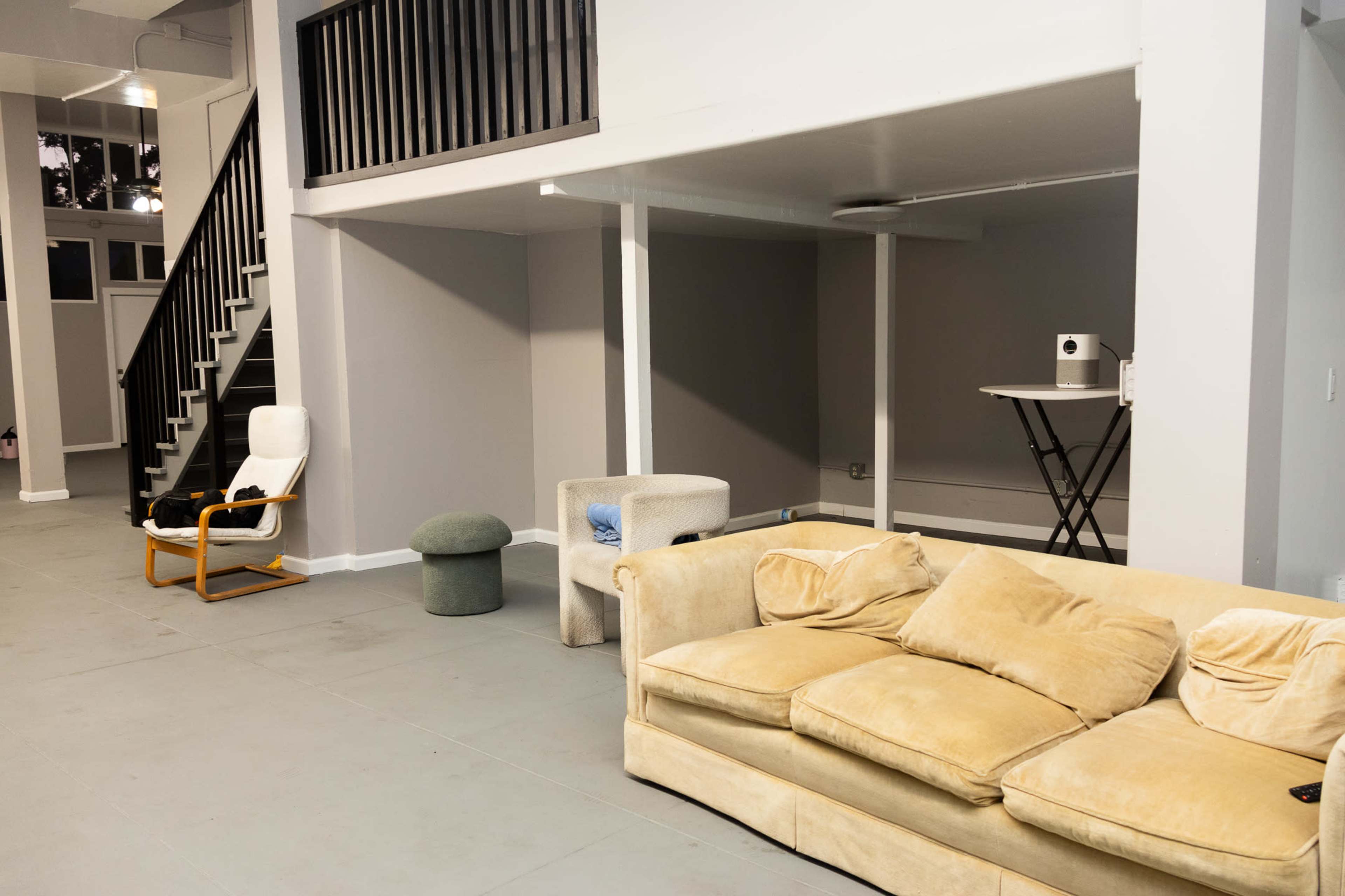 The image shows an open living space with a beige sofa, a white chair, a green pouf, and a small folding table near a staircase.