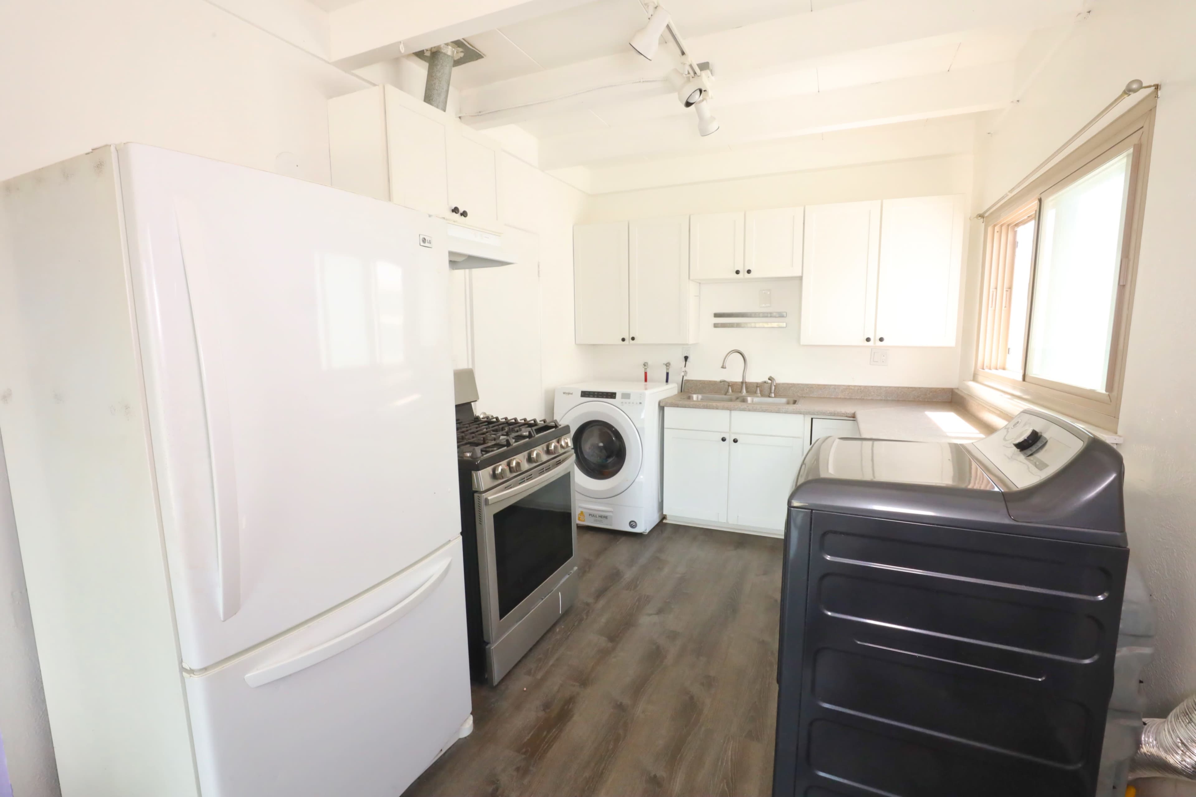 The image shows a modern kitchen with a white refrigerator, oven, washing machine, and dryer, along with white cabinets and a sink.