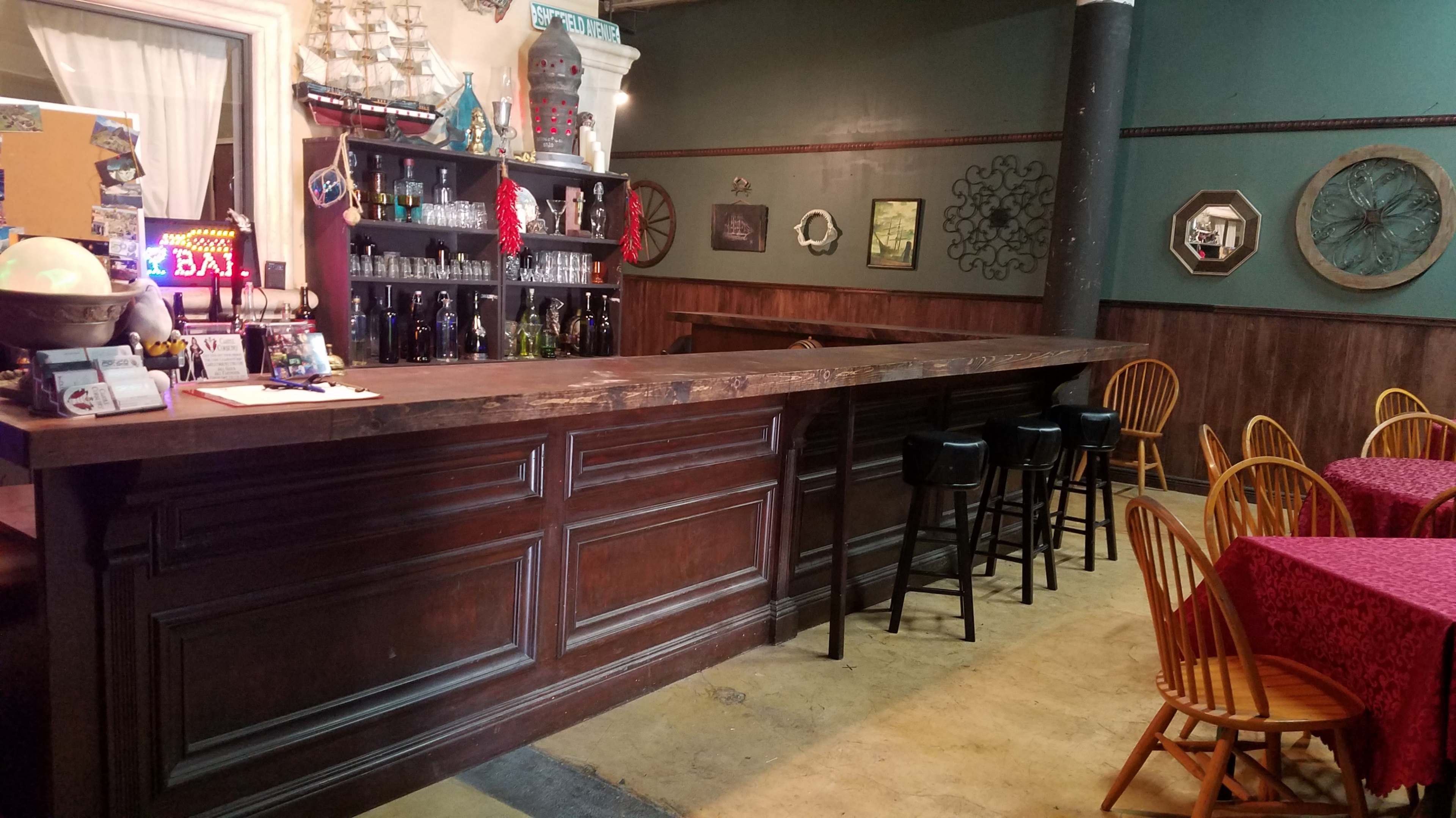 A wooden bar with black stools in front, surrounded by tables covered with red tablecloths in a rustic setting.