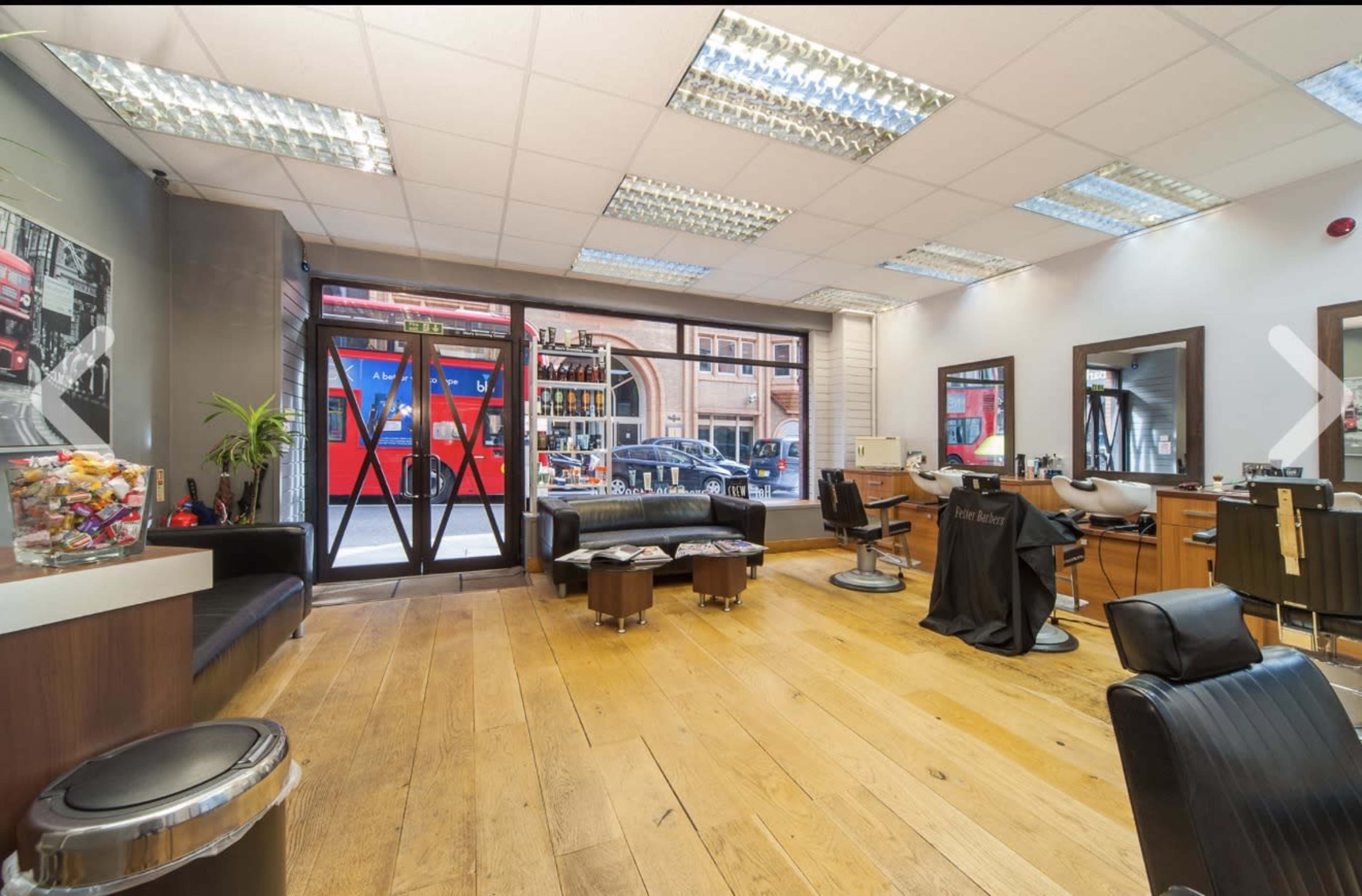 The interior of a hair salon features wooden flooring, several styling stations, and large windows displaying a street view with passing vehicles.