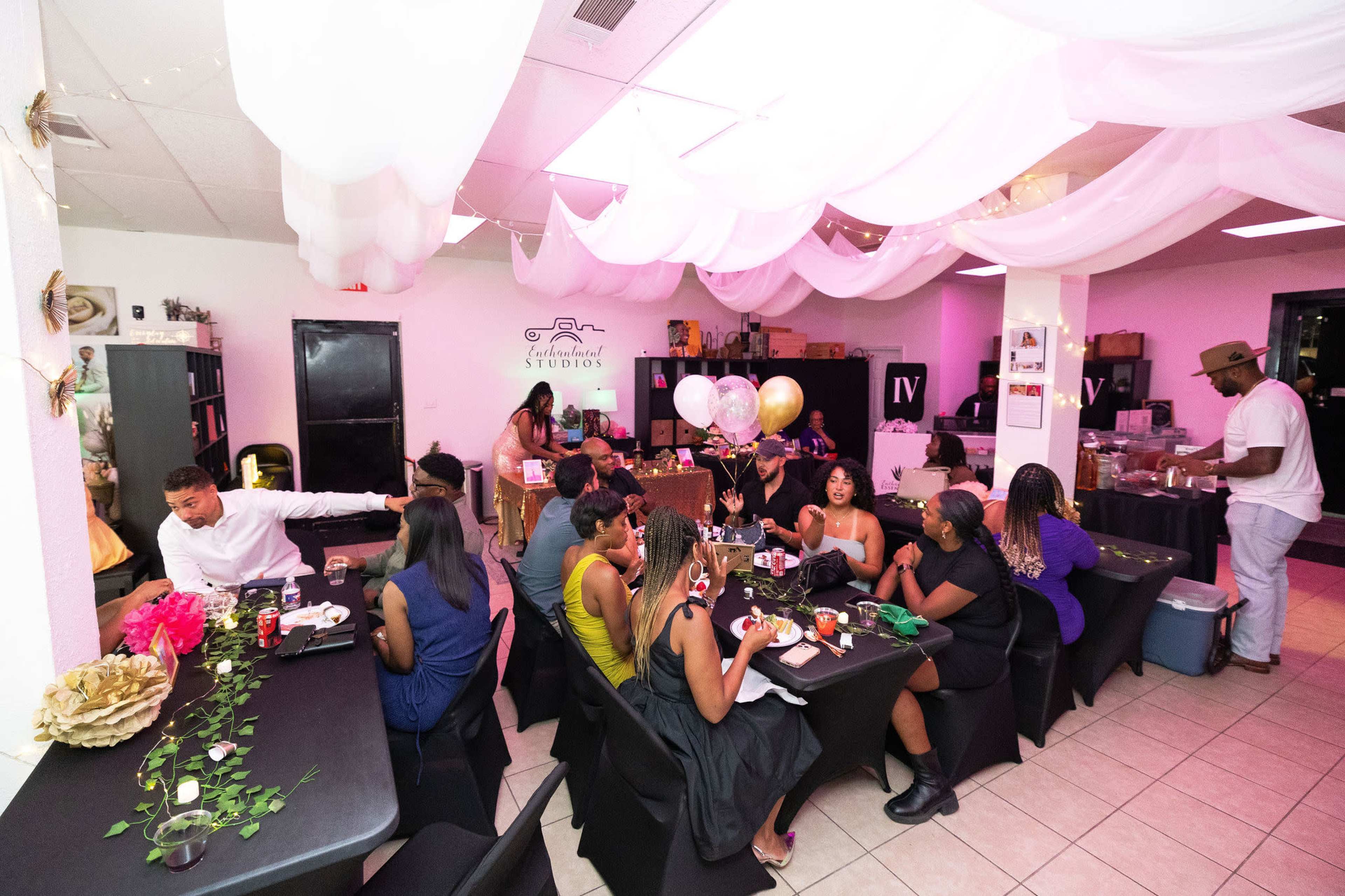 The image shows a lively indoor gathering with guests seated at tables, decorated with balloons and flowers, enjoying a meal in a well-lit space with pink lighting.