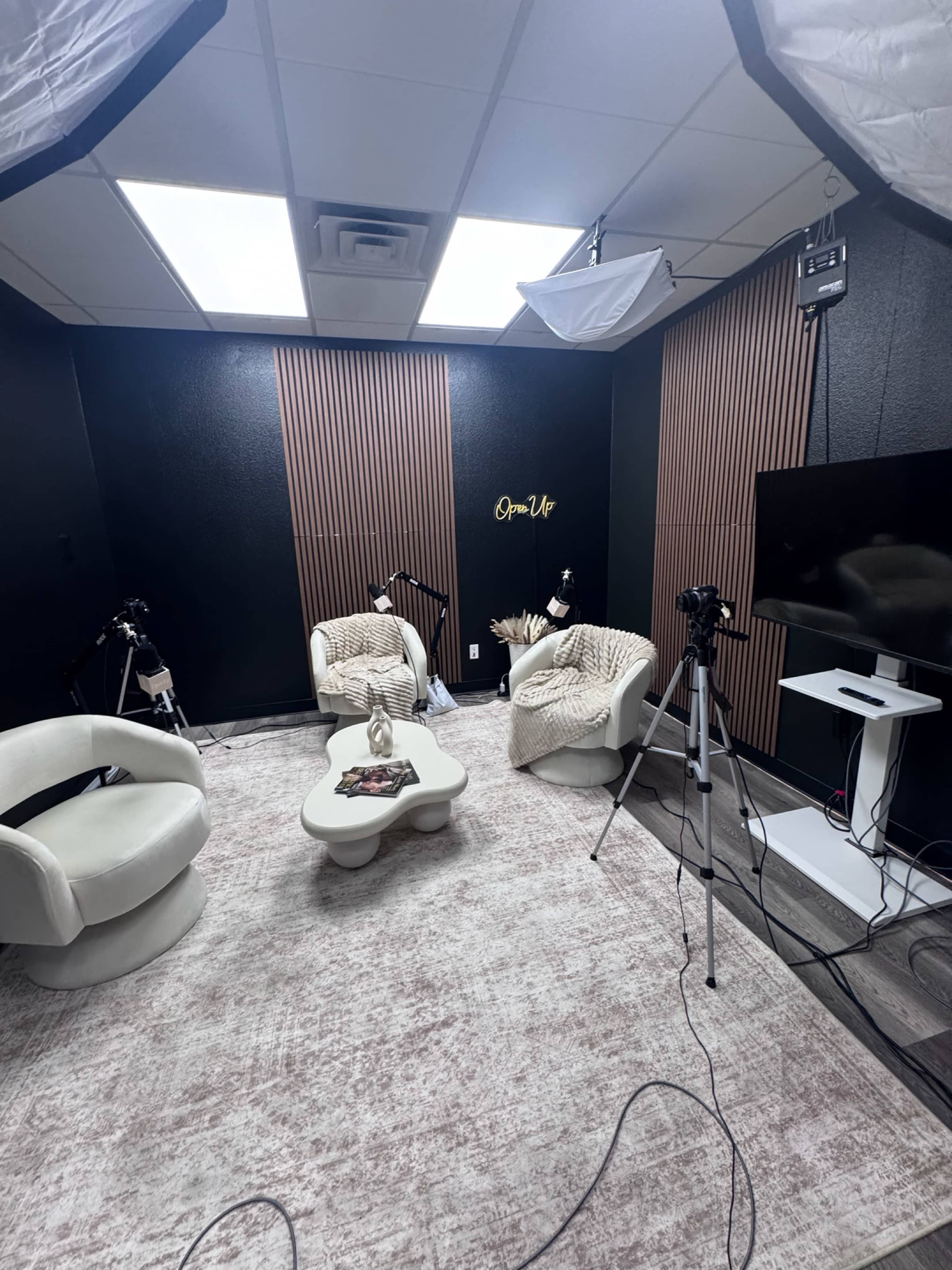 The image shows a modern, well-lit studio with two white seating chairs, a small coffee table, and videography equipment set up against a black and wood-paneled wall.