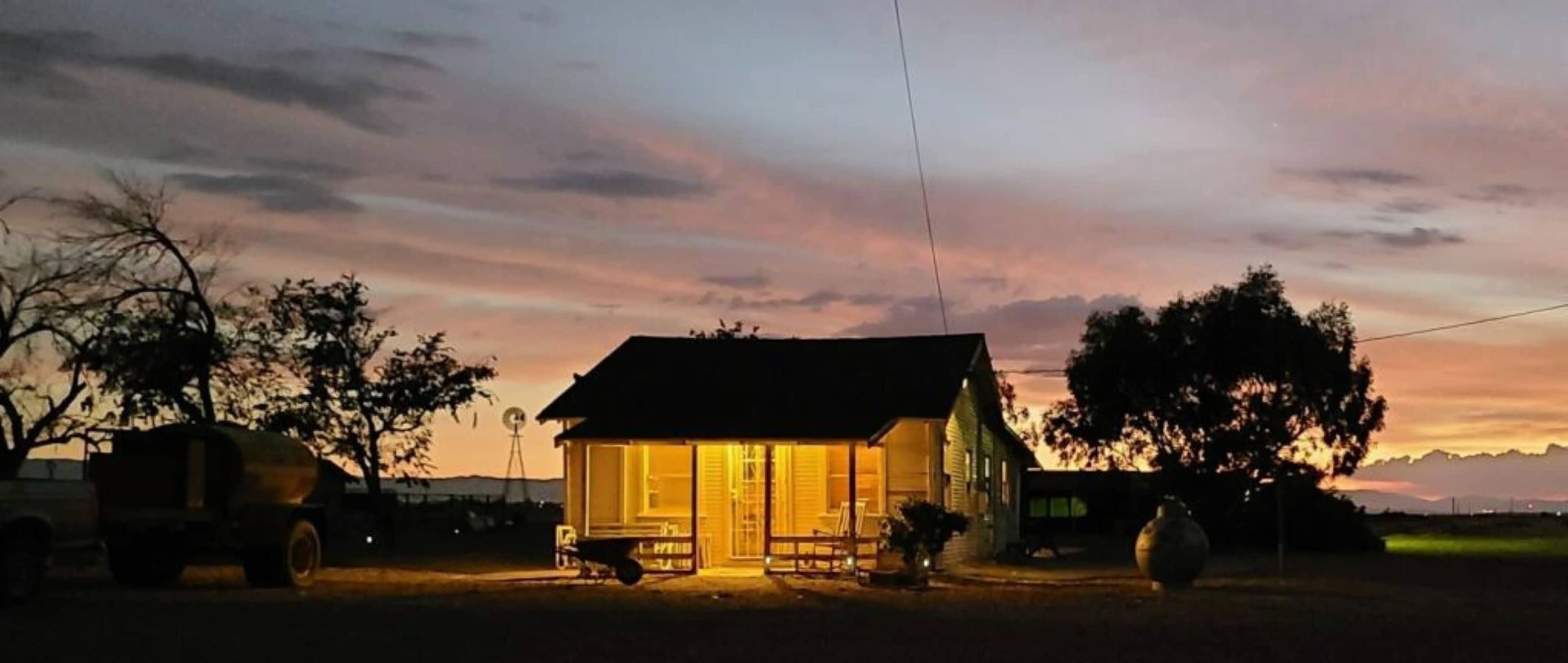 A small house with illuminated windows sits against a colorful sunset, surrounded by trees and a silo in the distance.