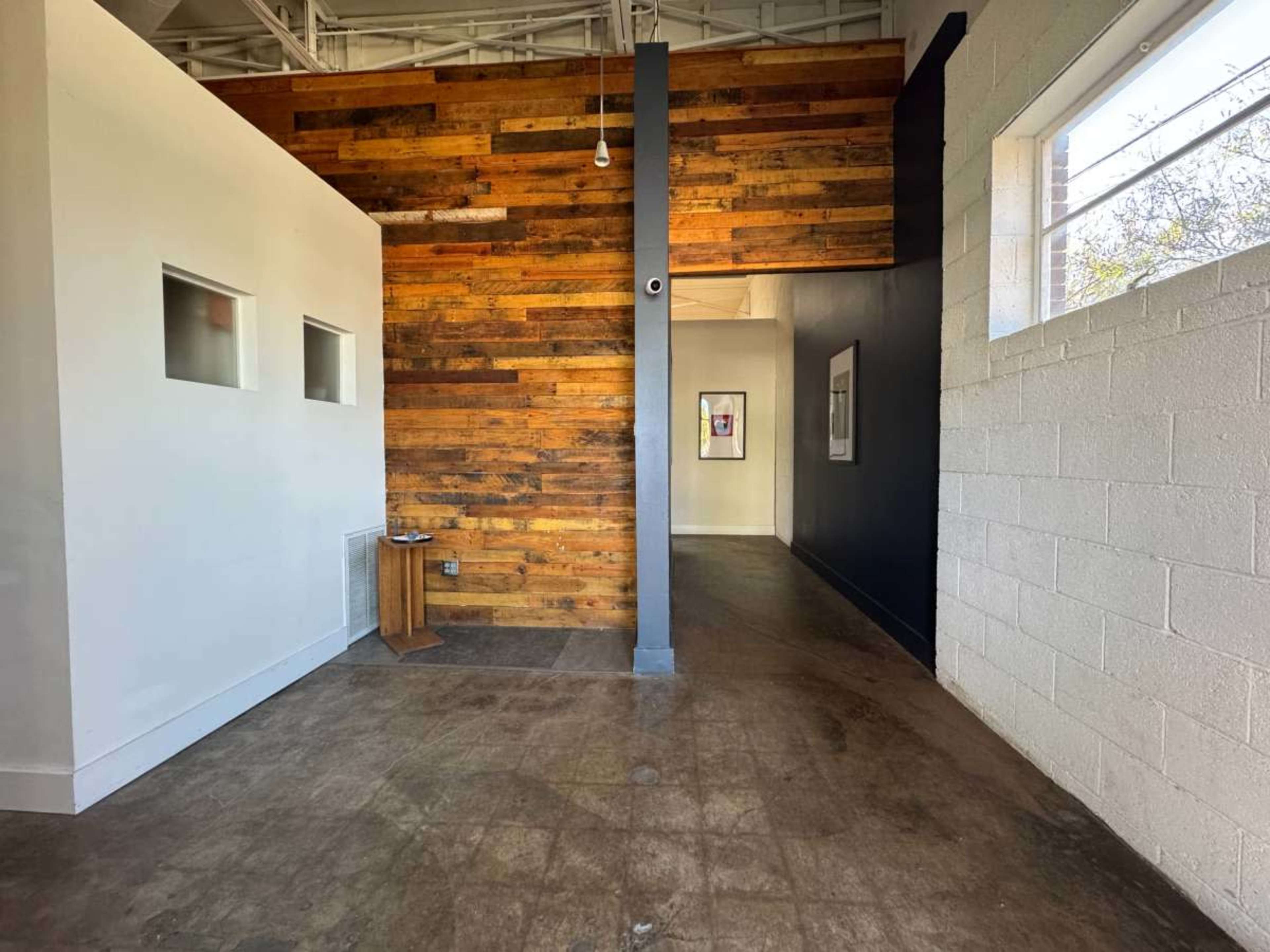 The image shows a room with a textured wooden accent wall, leading into a hallway with artworks displayed on the walls and large windows allowing natural light.