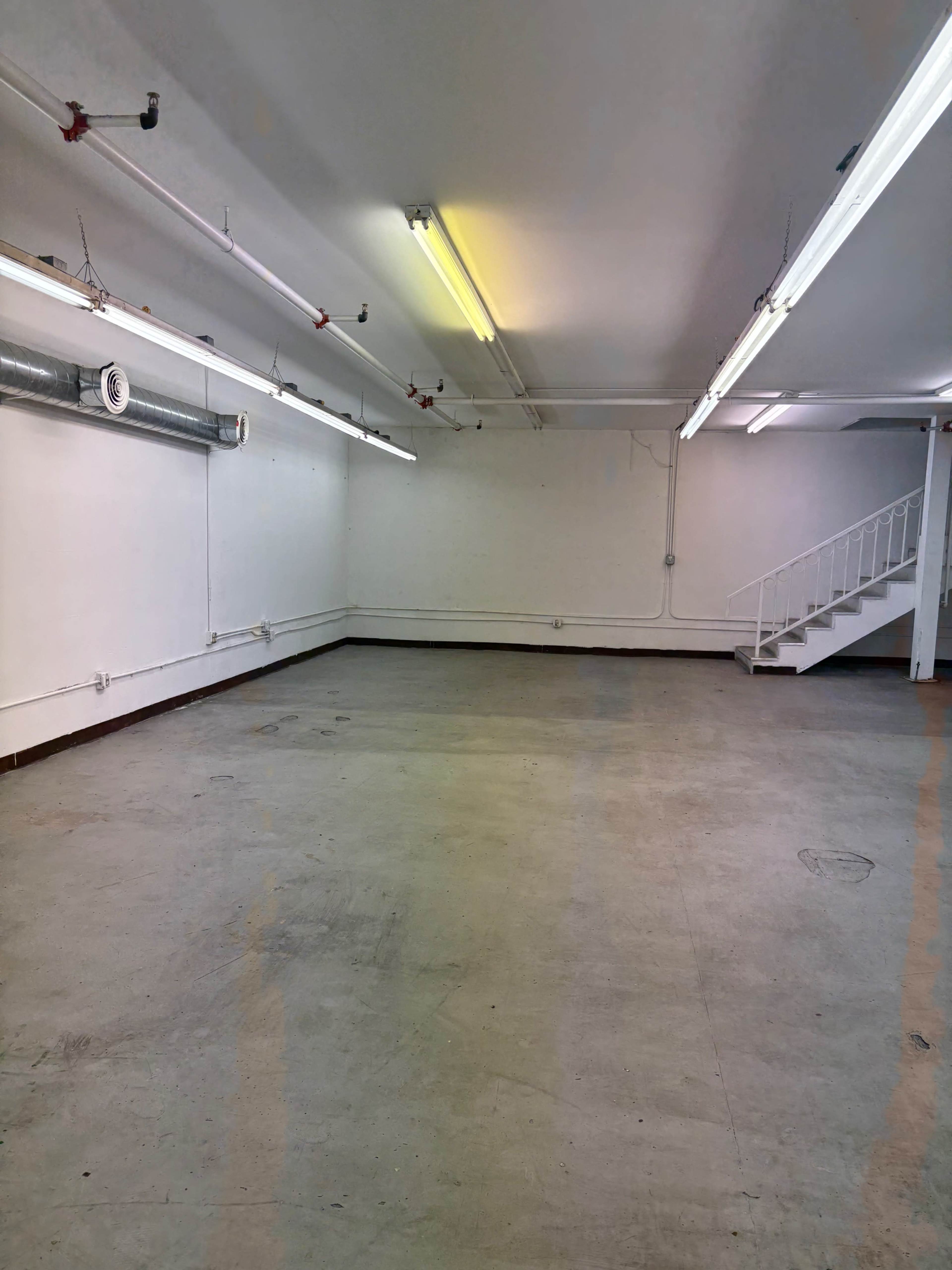 The image shows a large, empty room with concrete flooring, white walls, and overhead fluorescent lights, along with a staircase in the back corner.
