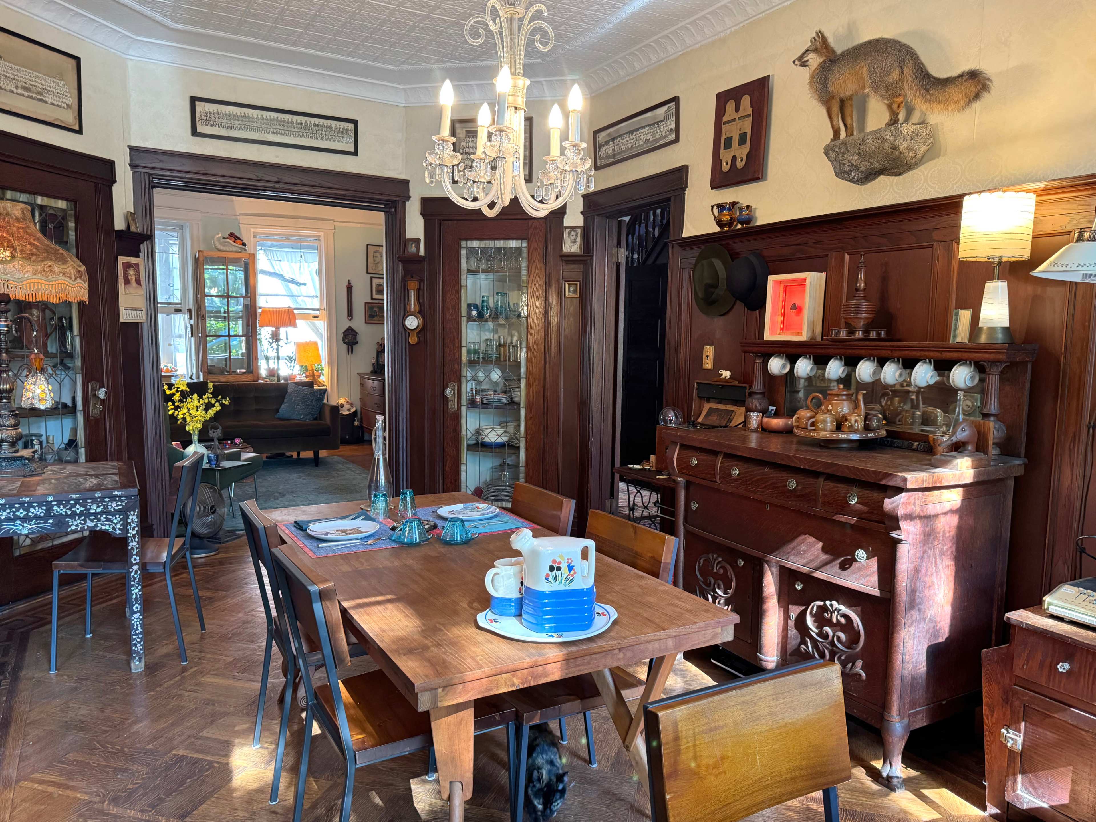 The image shows a warmly lit dining area with a wooden table set for a meal, surrounded by vintage decor and a display cabinet filled with glassware.