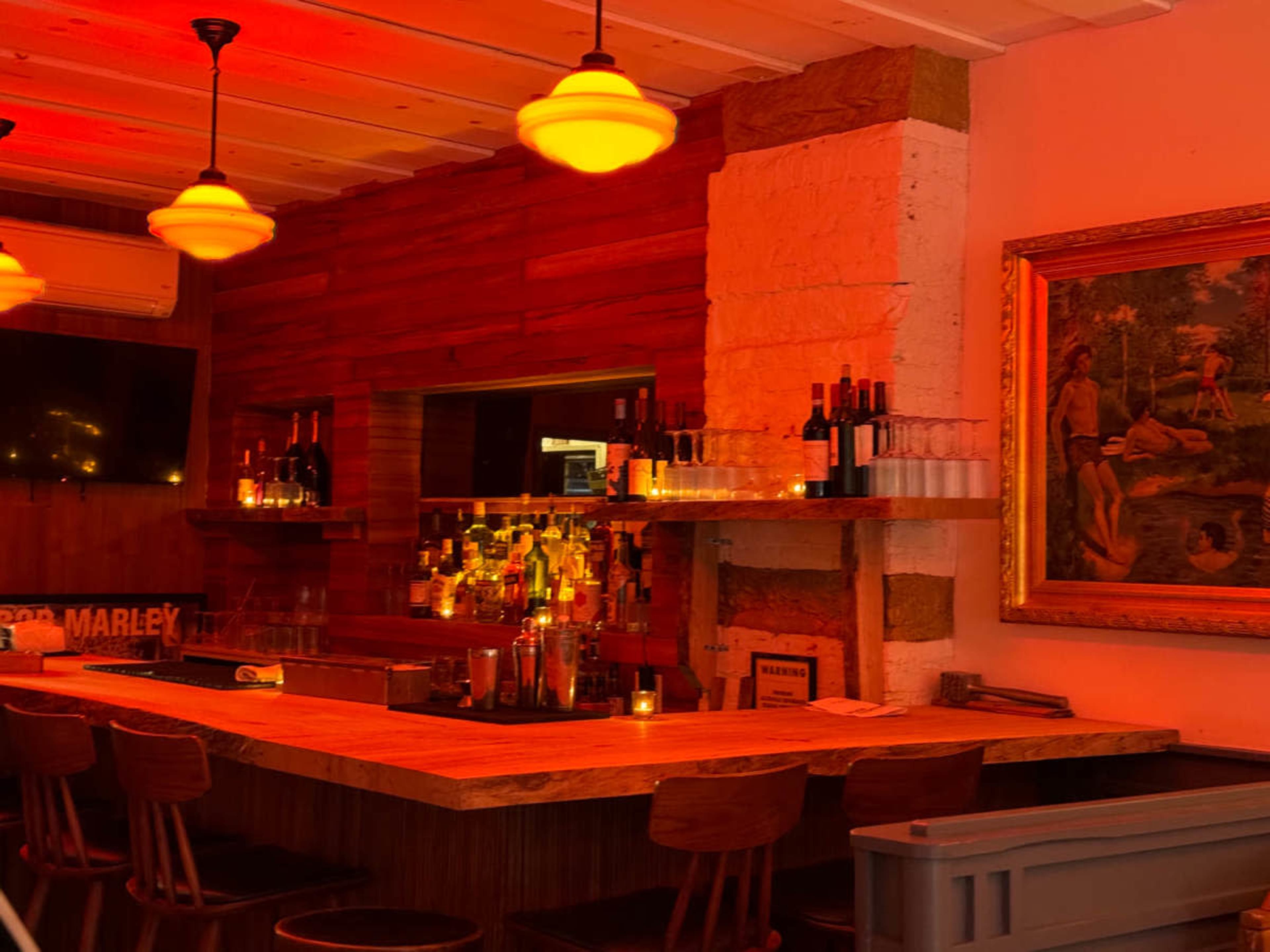 The image shows a dimly lit bar with a wooden counter, shelves filled with bottles, and decorative artwork on the wall.