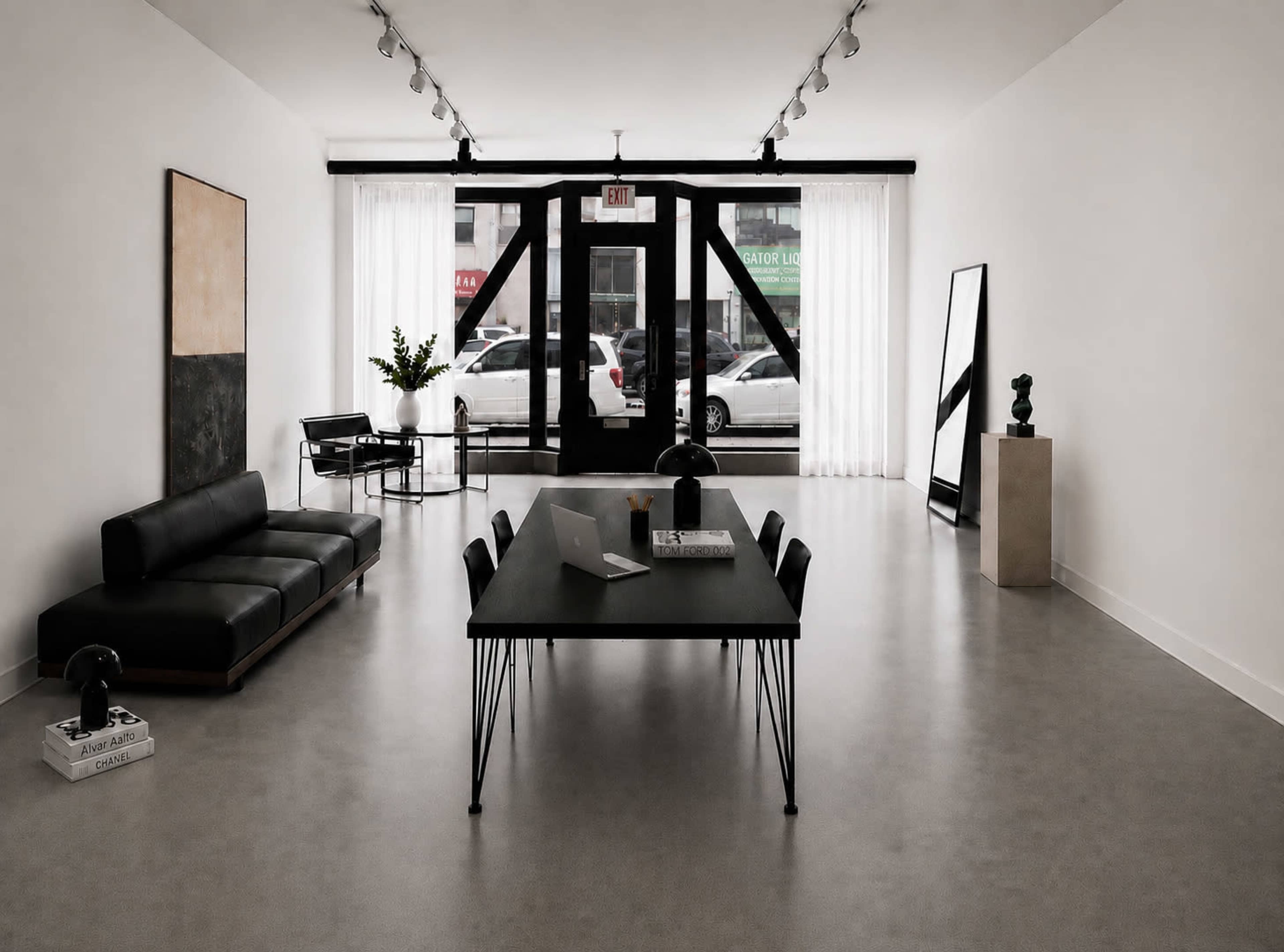 A modern interior space features a sleek black sofa, a minimalist dining table with chairs, and large windows along one wall that overlook a street.