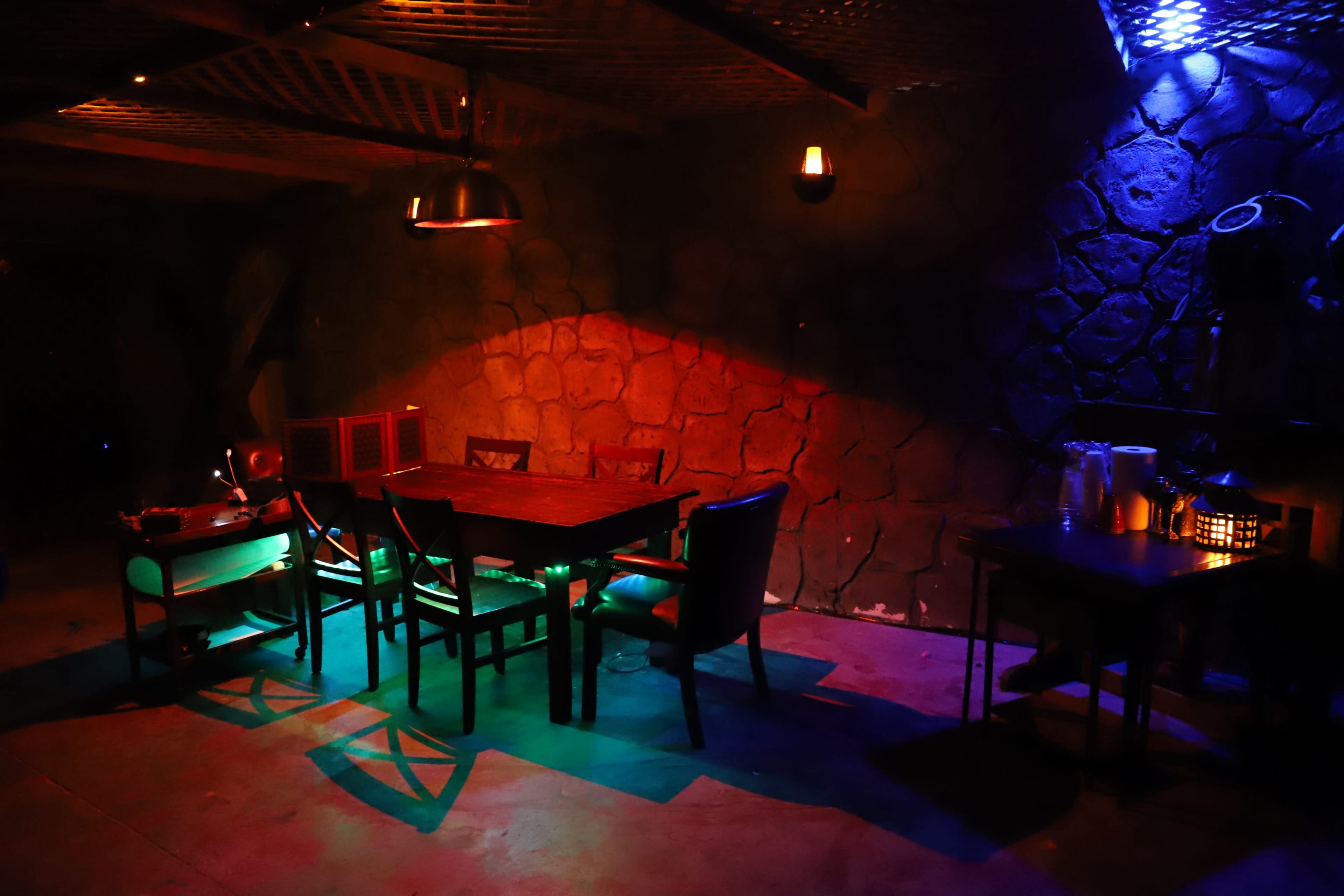 A dimly lit dining area features a large wooden table surrounded by chairs, with colorful lights casting shadows on the stone walls.