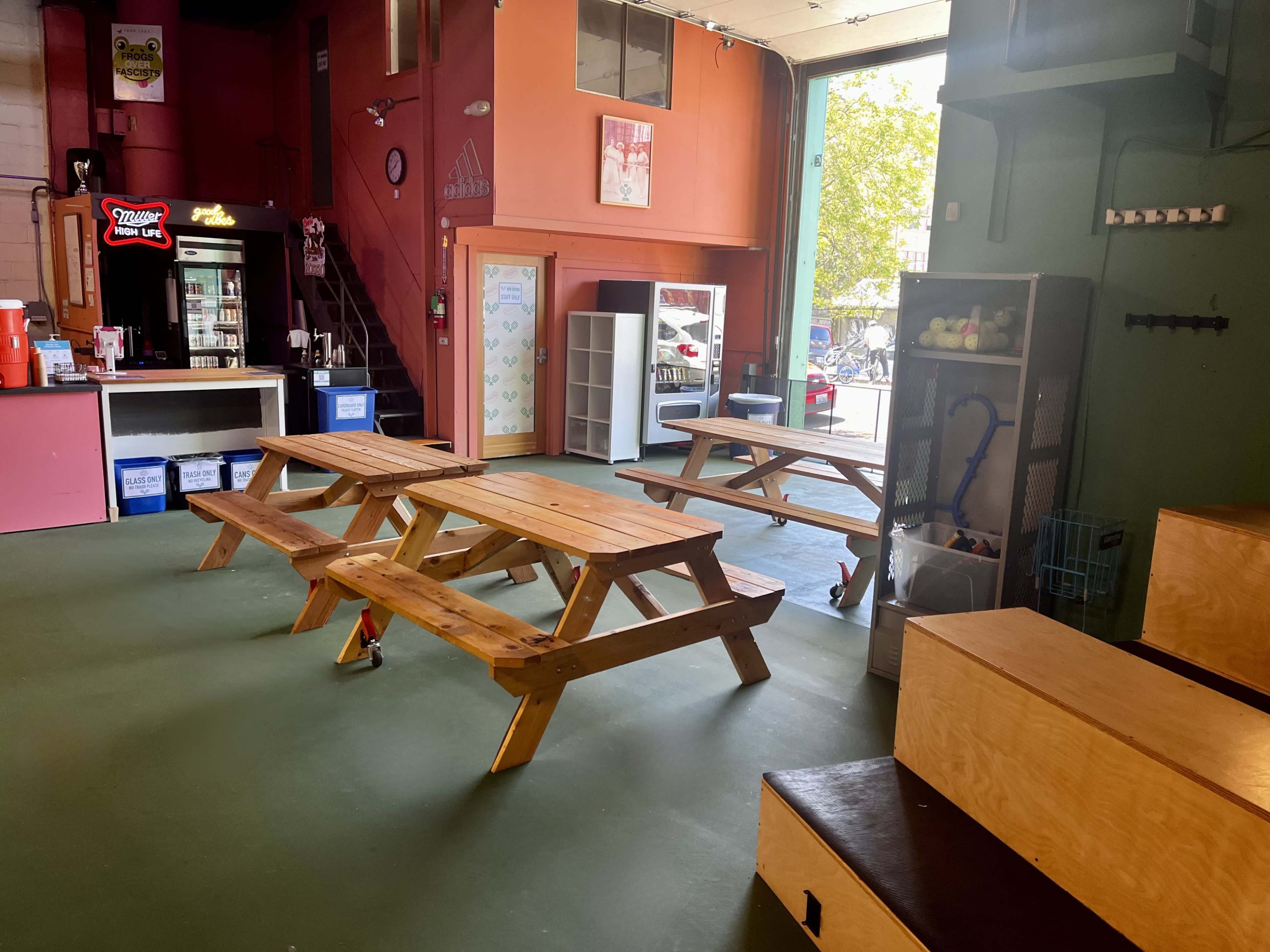 The image shows an indoor space with wooden picnic tables, benches, and a service counter in a colorful setting.