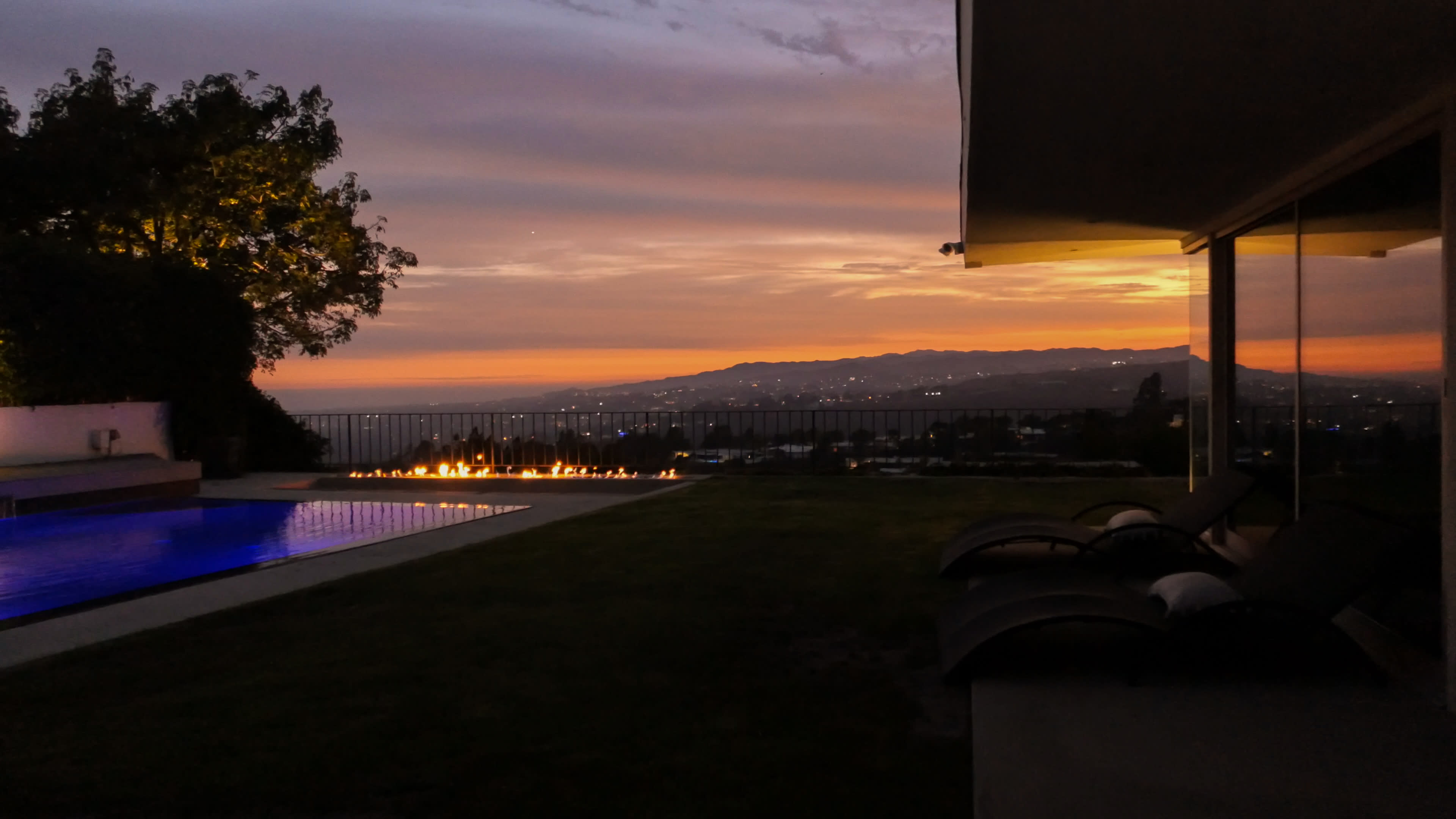 The image shows a sunset view over a pool with lounge chairs, framed by a tree and distant mountains.