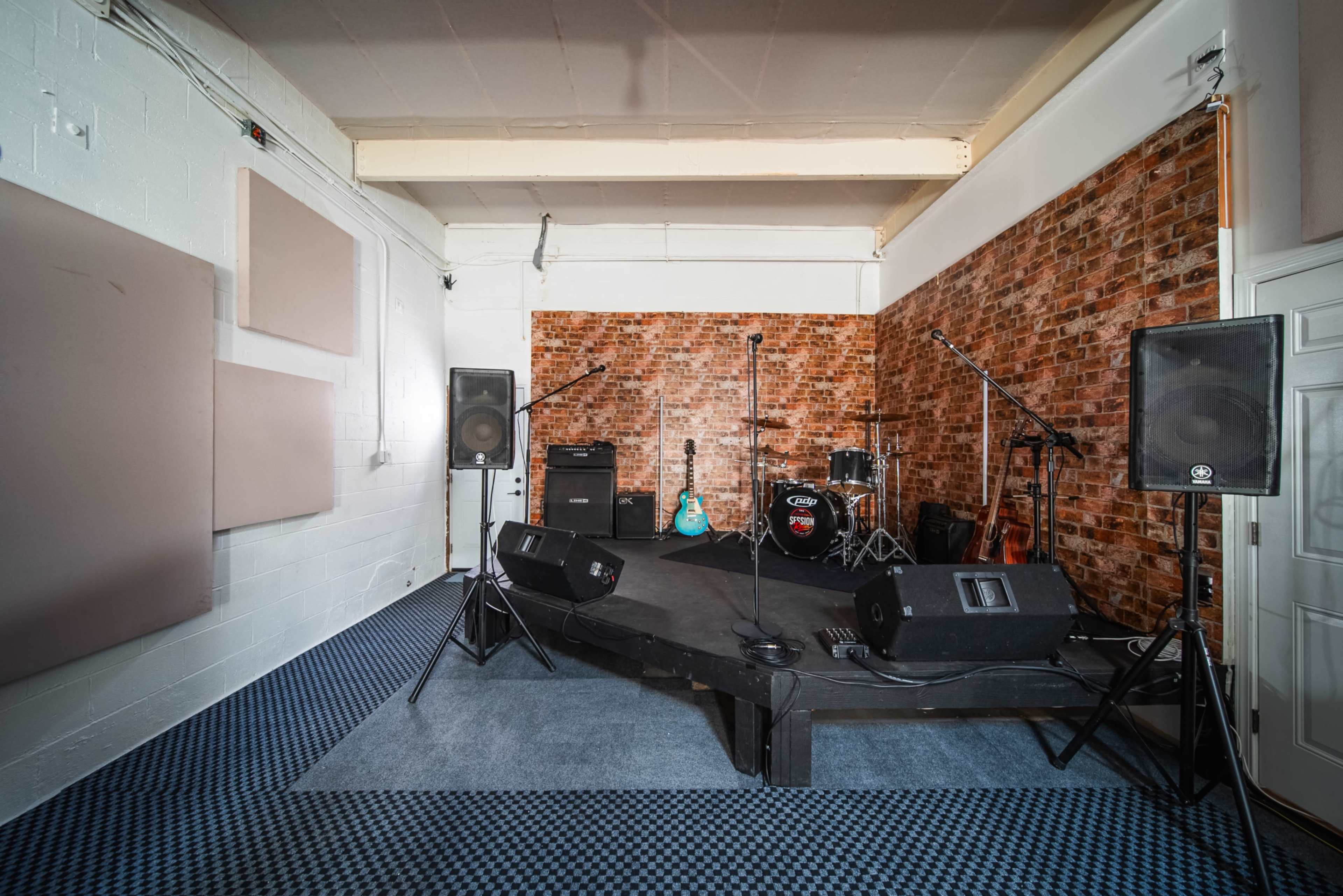 The image shows a small music performance stage with amplifiers, microphones, and a drum set against a brick wall backdrop.