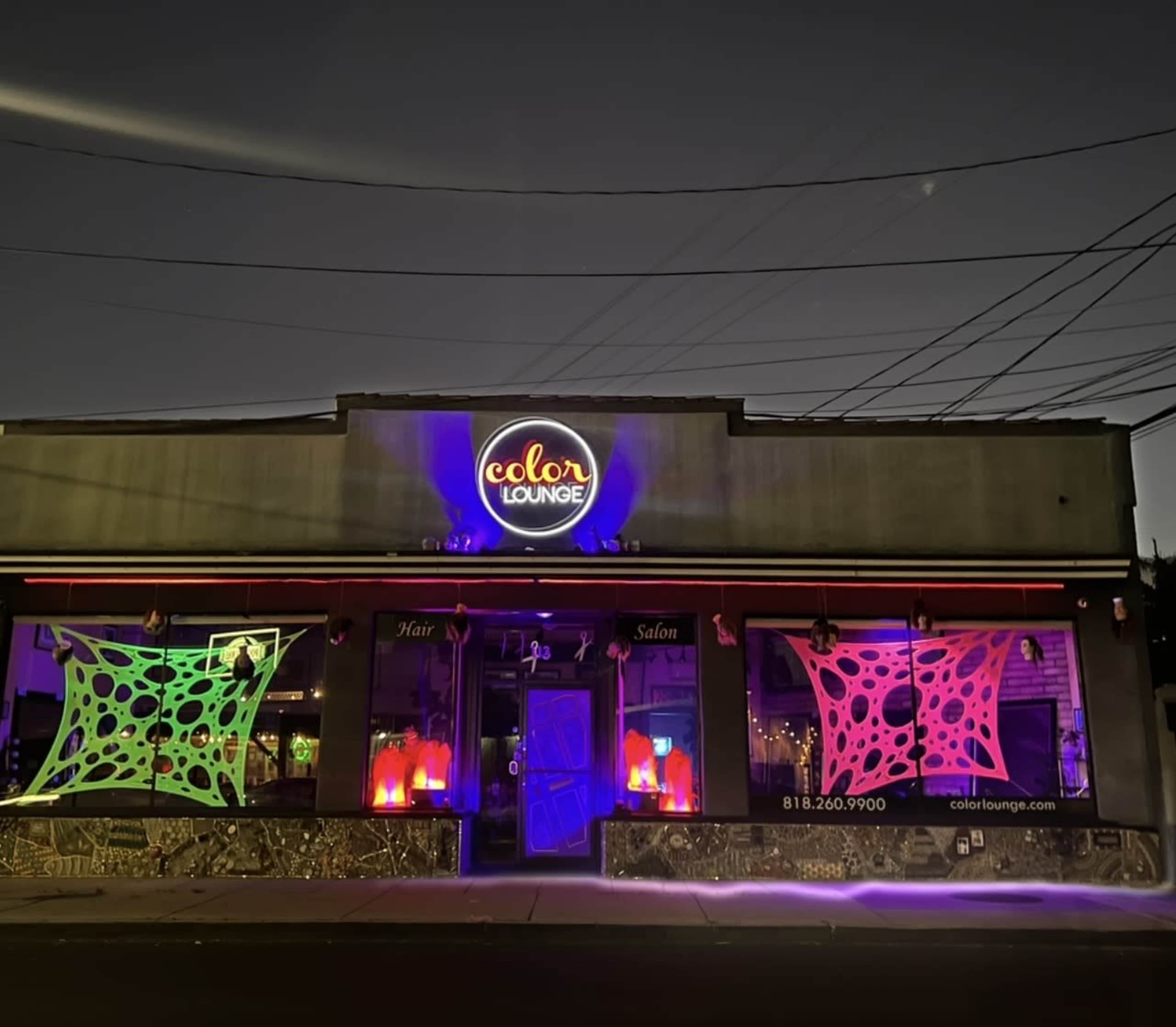 The image shows a hair salon called "Color Lounge" with colorful neon lights and spider web decorations displayed in the windows at night.
