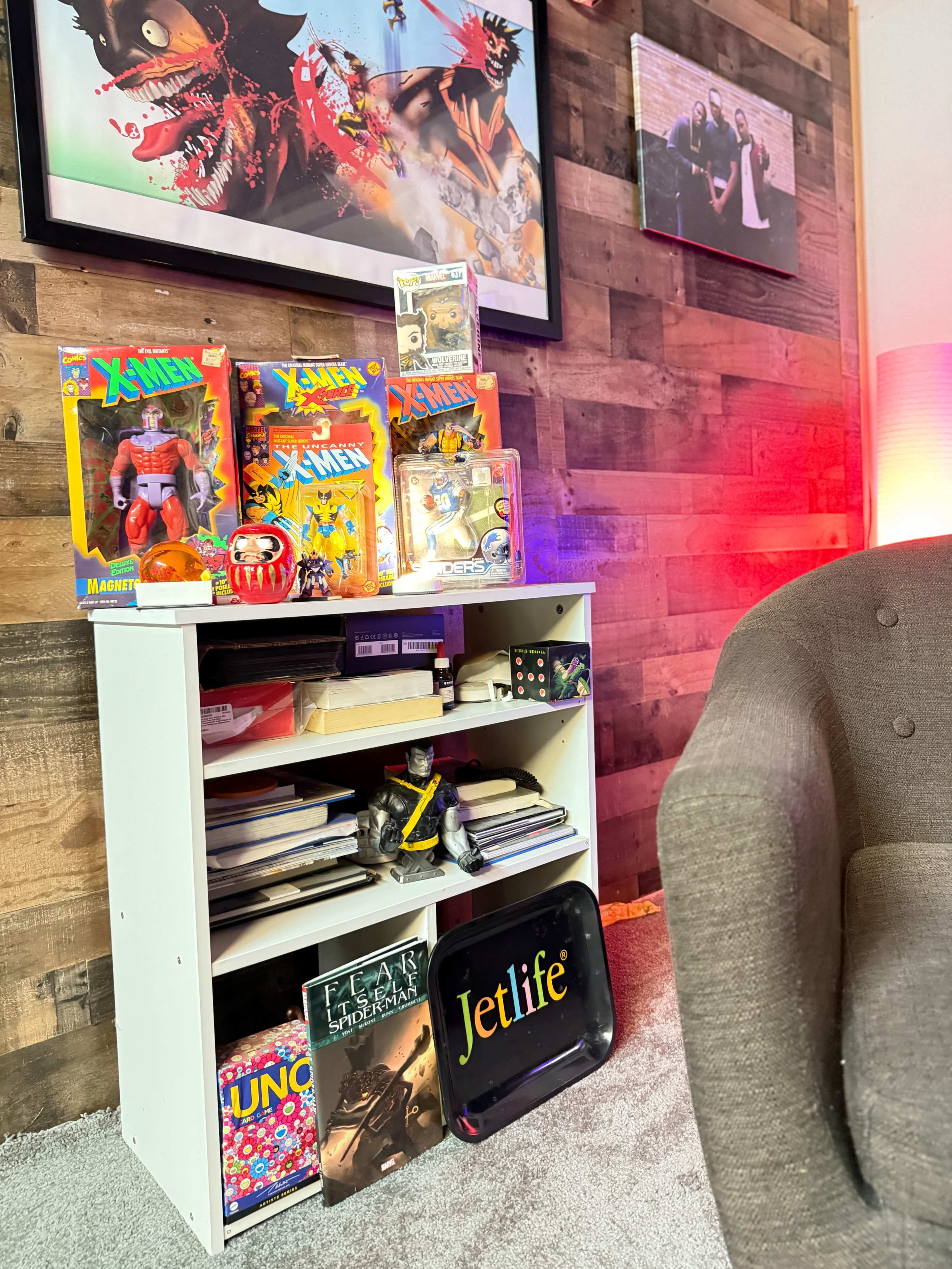 A white bookshelf contains various toys, books, and collectibles, set against a textured wooden wall and illuminated by colorful lighting.