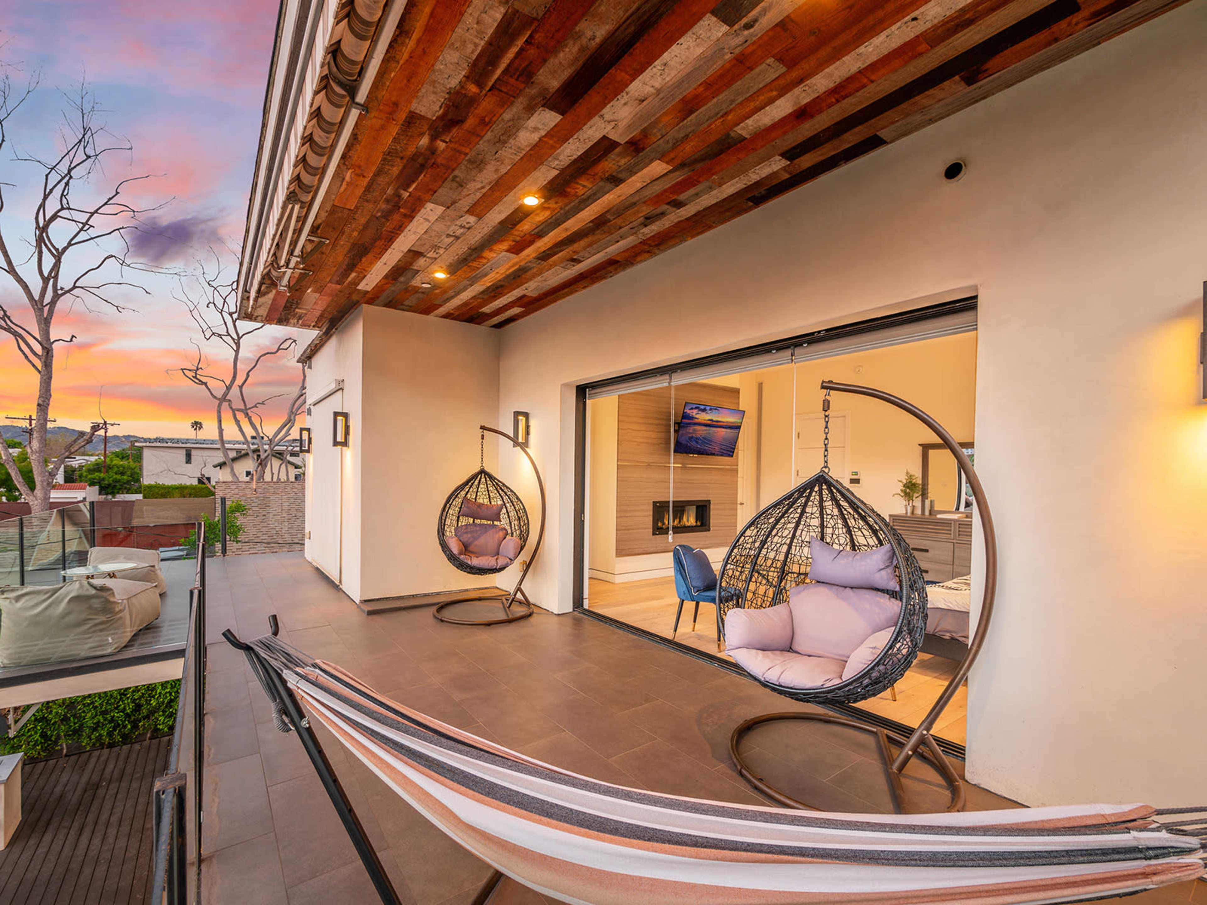 A modern patio features two hanging chairs and a hammock, with a view of a sunset sky and an interior room visible through large glass doors.