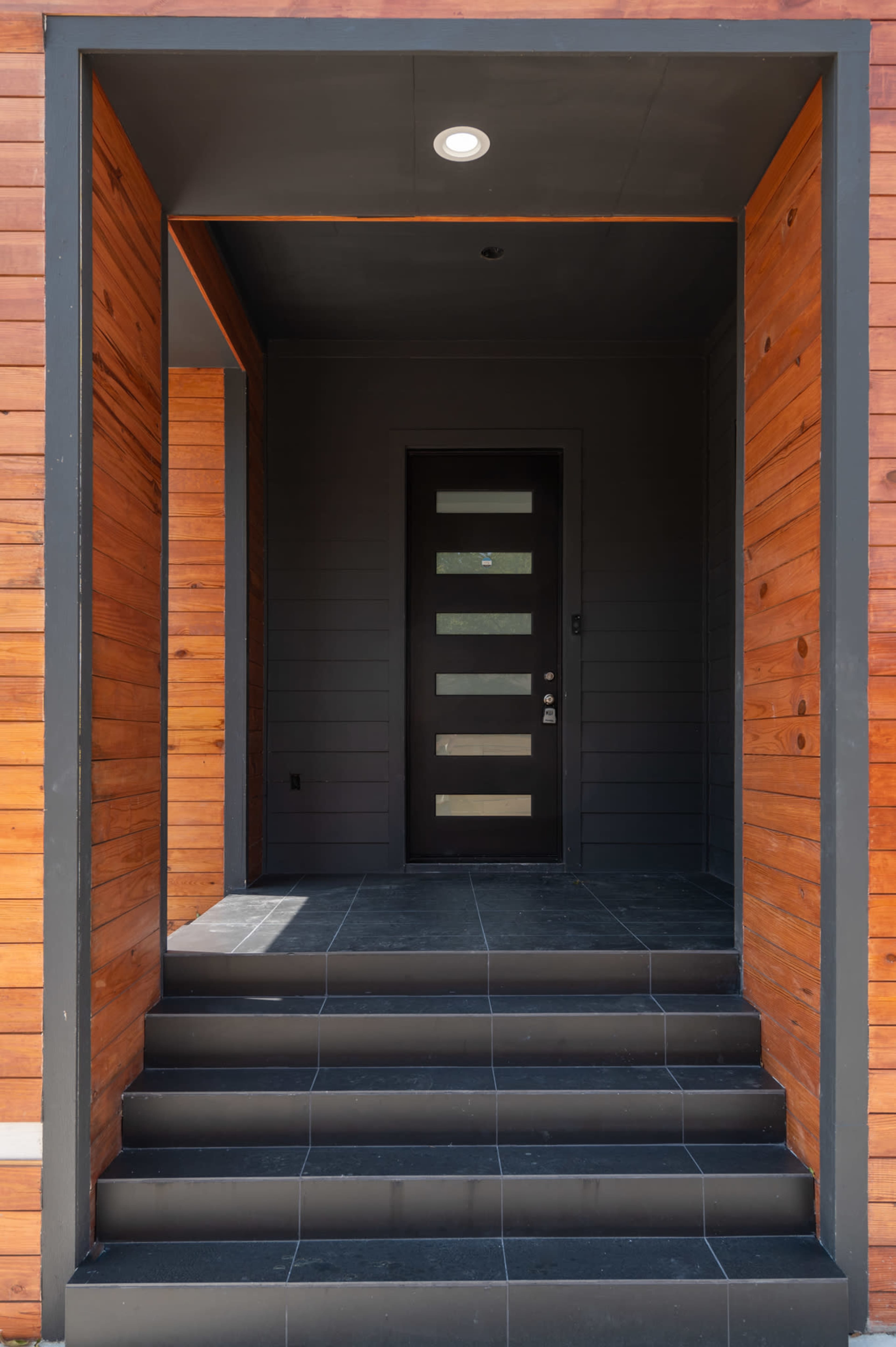 The image shows a modern entryway with wooden siding, a black front door, and tiled steps leading up to it.