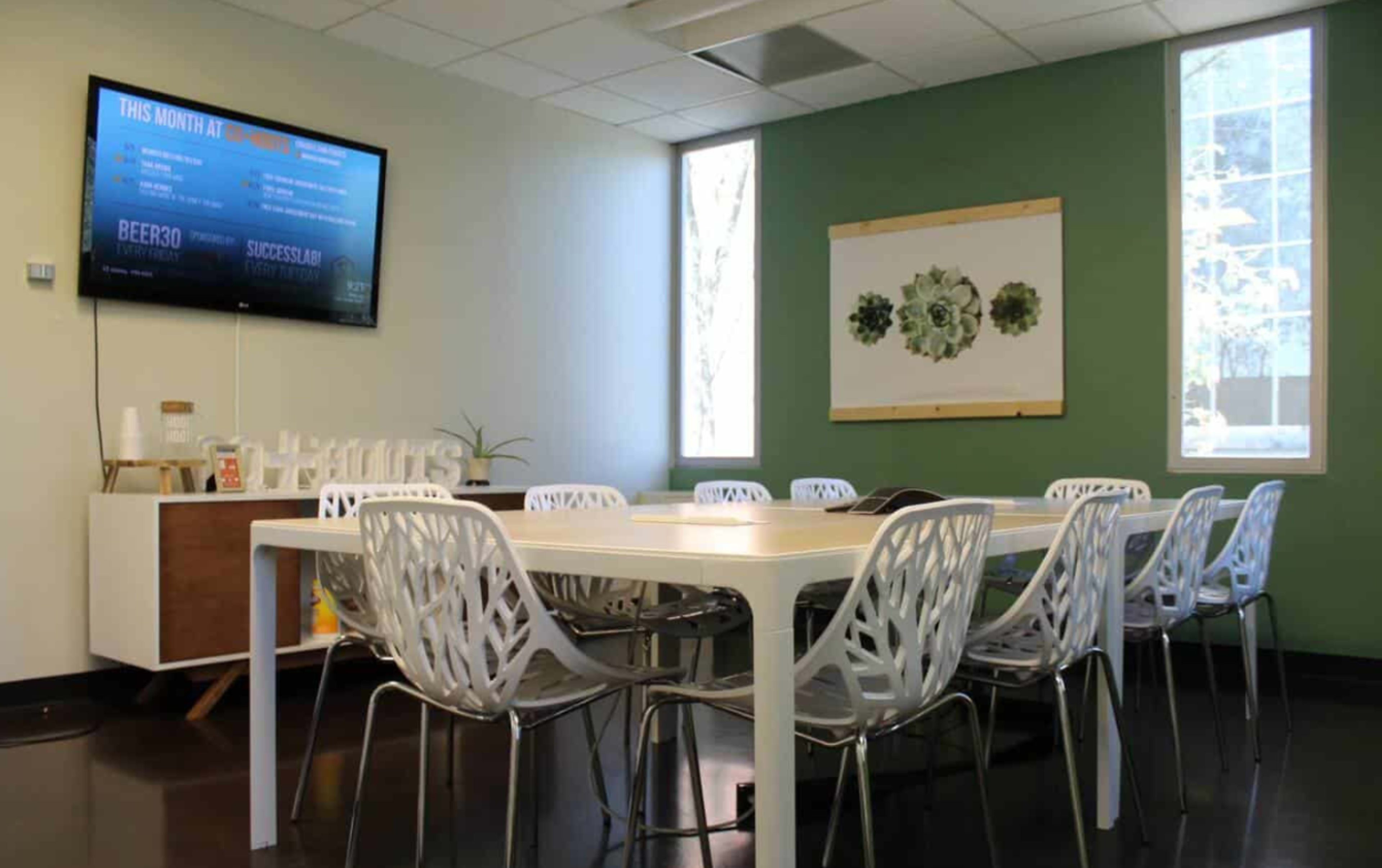 The image shows a modern meeting room with a large table surrounded by white chairs, a television mounted on the wall, and natural light coming through two windows.