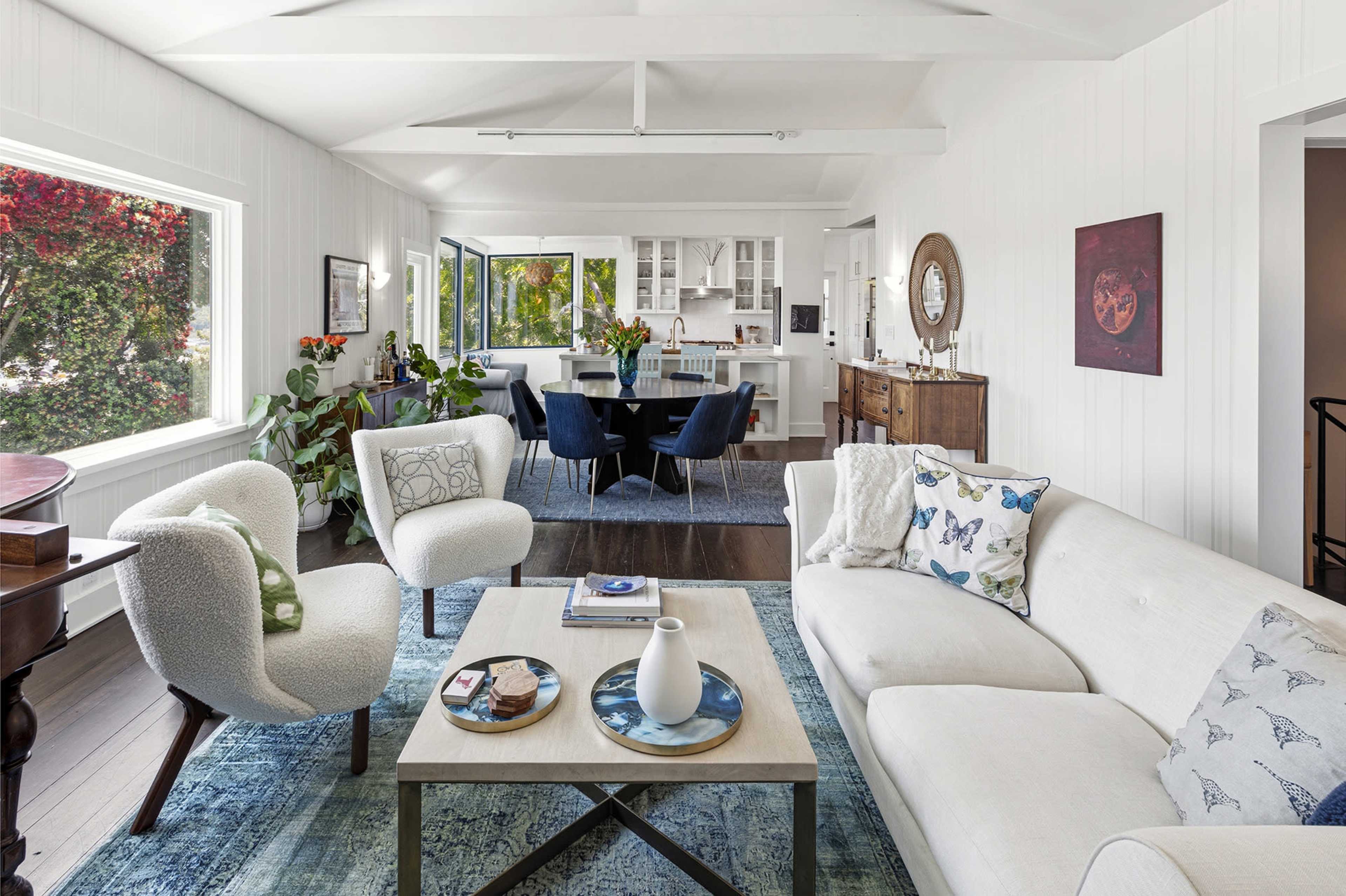 A bright living room features a light-colored sofa, decorative armchairs, and a coffee table, all set against a backdrop of large windows overlooking greenery.