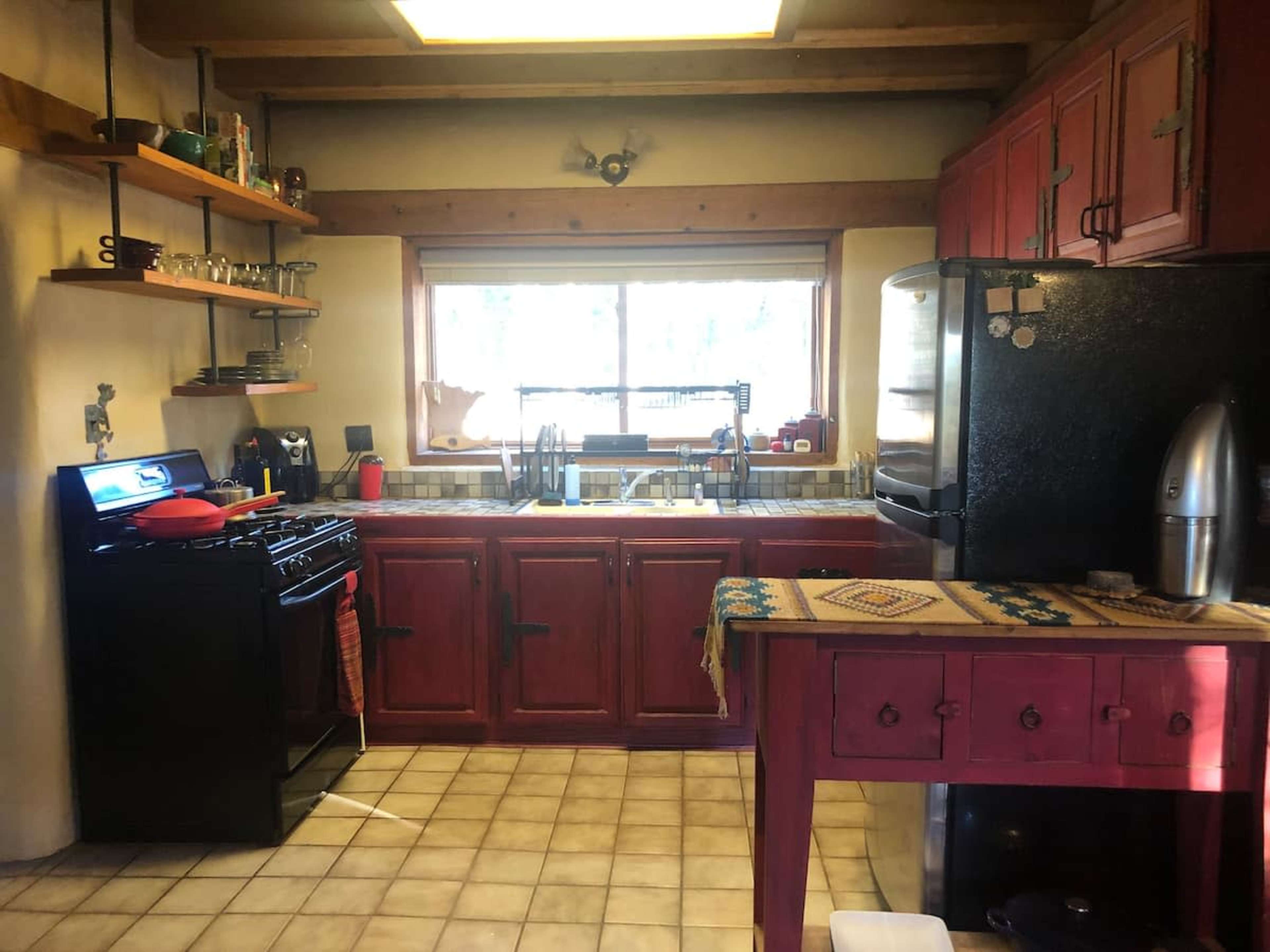 A rustic kitchen features red cabinetry, a gas stove, a large window, and a black refrigerator, with a table and shelves displaying various kitchen items.