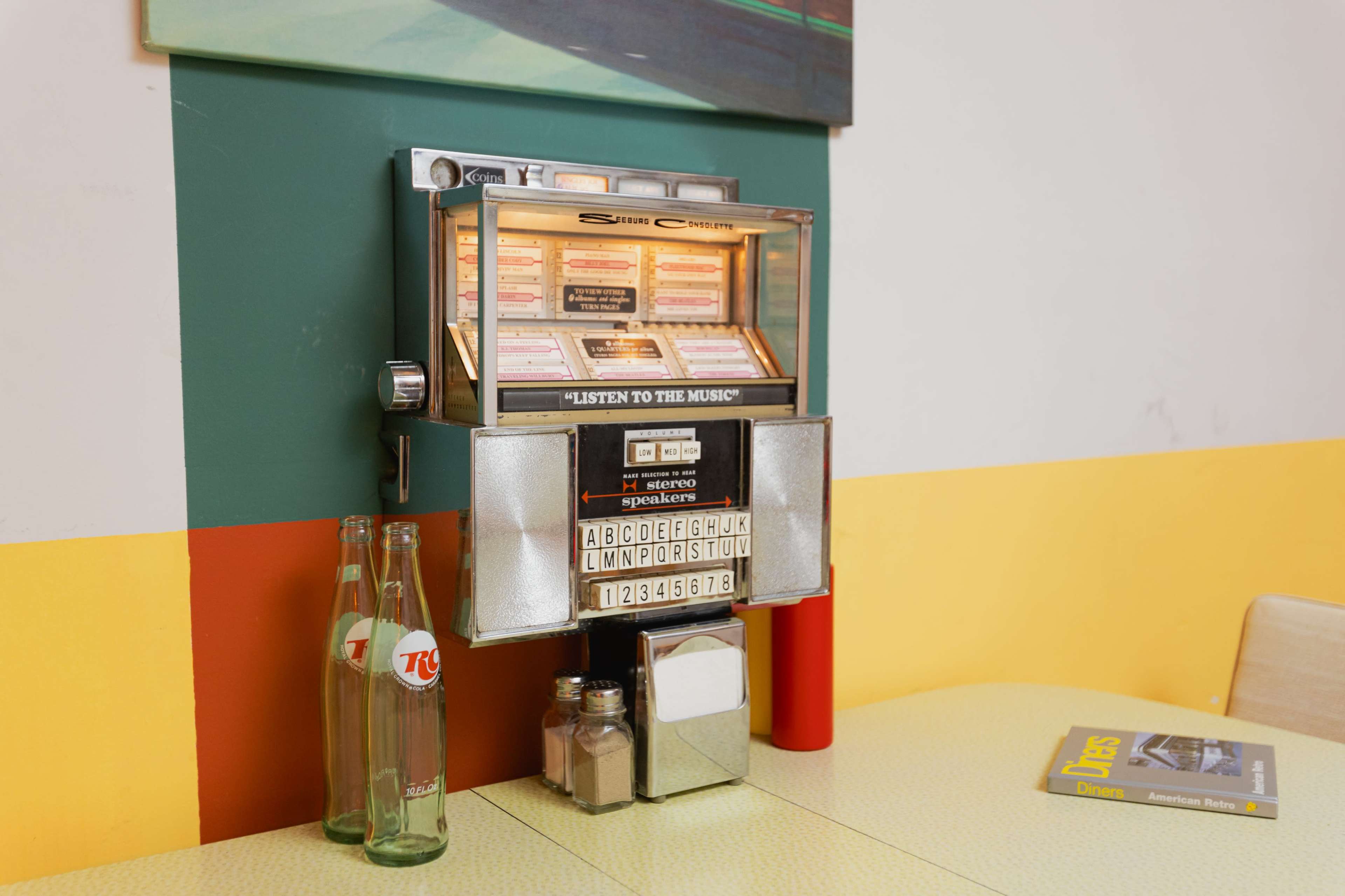 A vintage jukebox with glass panels and visible selection buttons is mounted on a colorful wall in a diner-style setting.