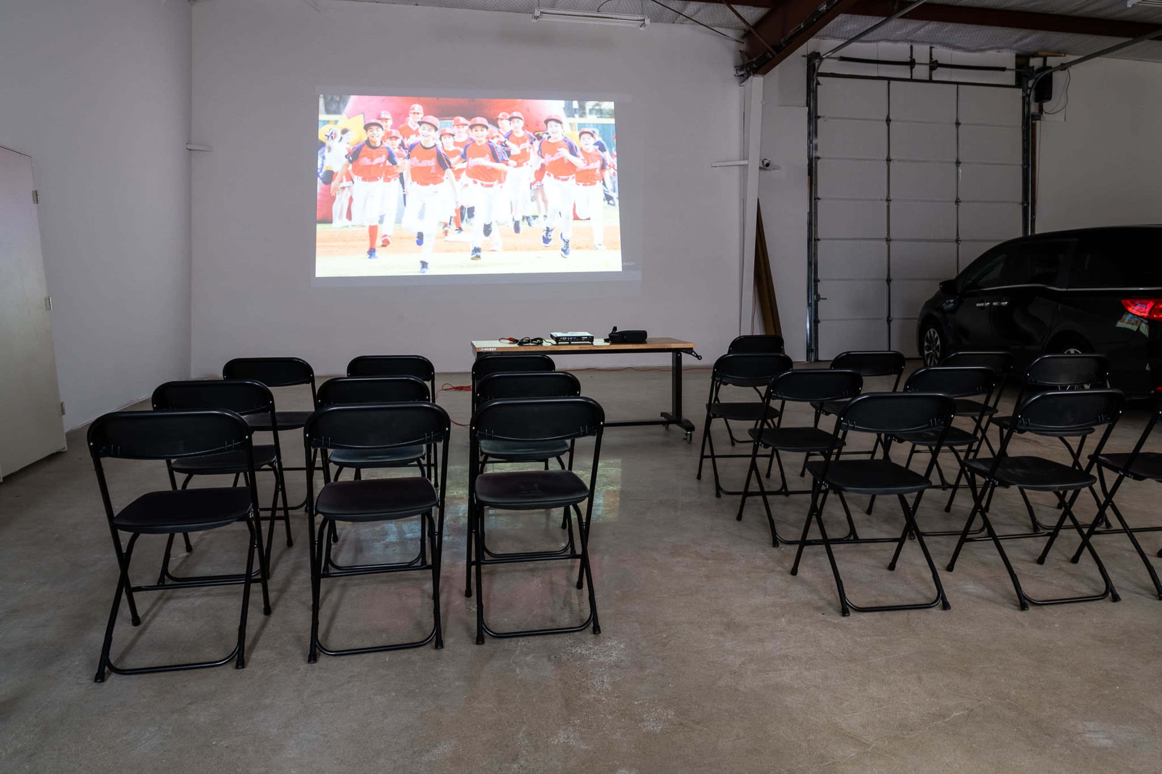 A row of black folding chairs faces a projector screen displaying a group of people in matching outfits, set up in a bare, well-lit room.