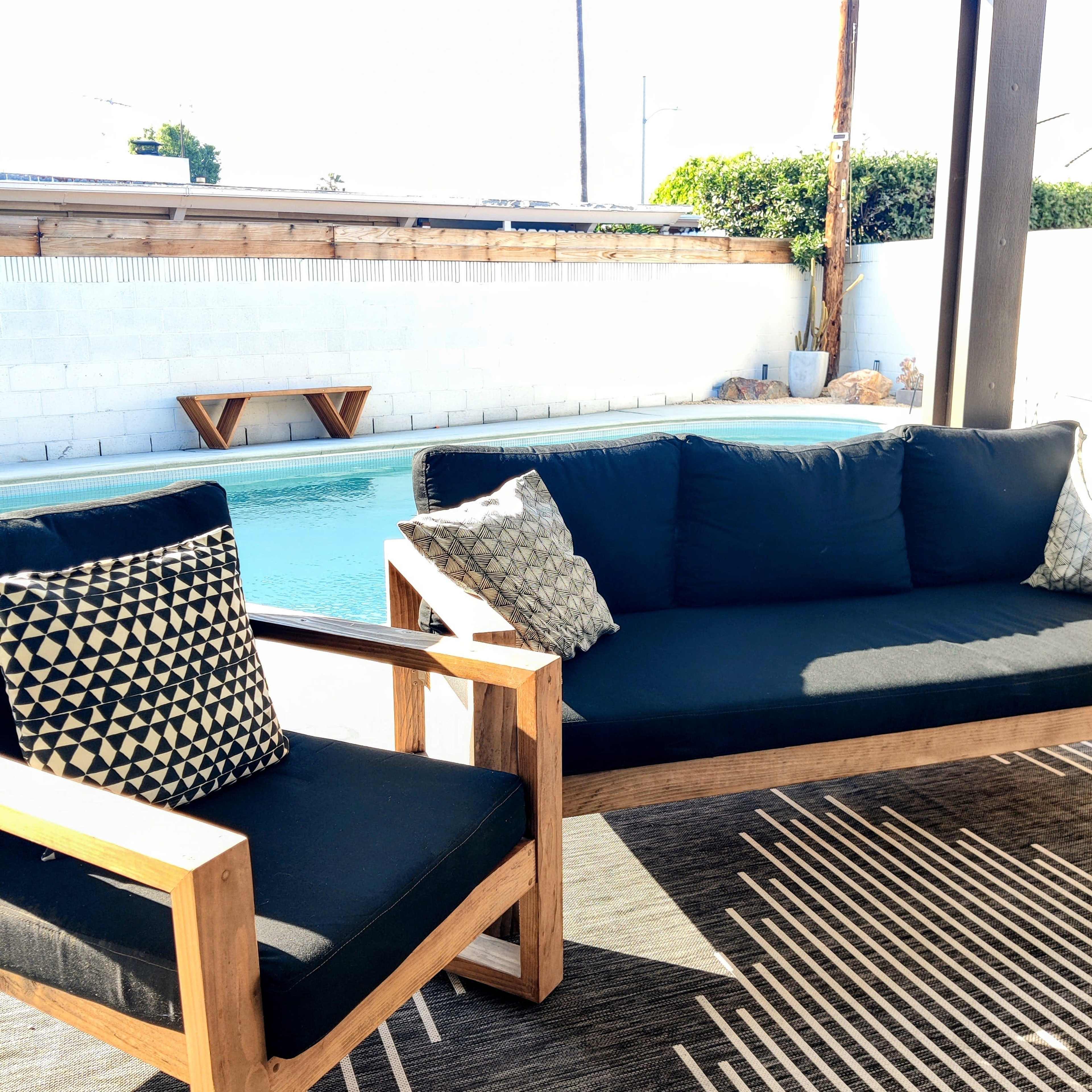 A wooden patio set with dark cushions and patterned pillows is positioned beside a swimming pool surrounded by a white wall.
