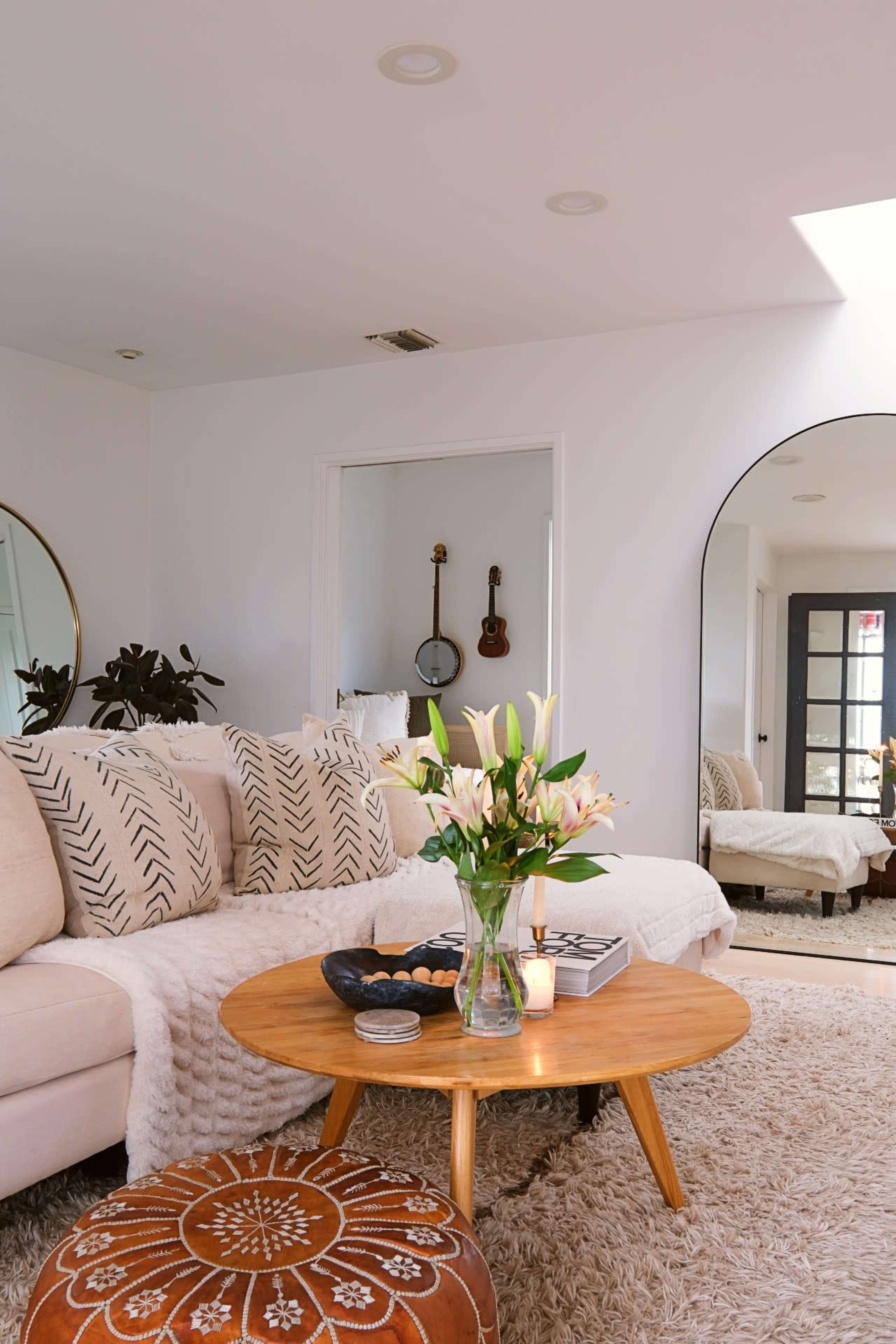 A cozy living room features a large beige sofa with decorative pillows, a wooden coffee table with a vase of flowers, and a round pouf, all set against a bright and airy backdrop.