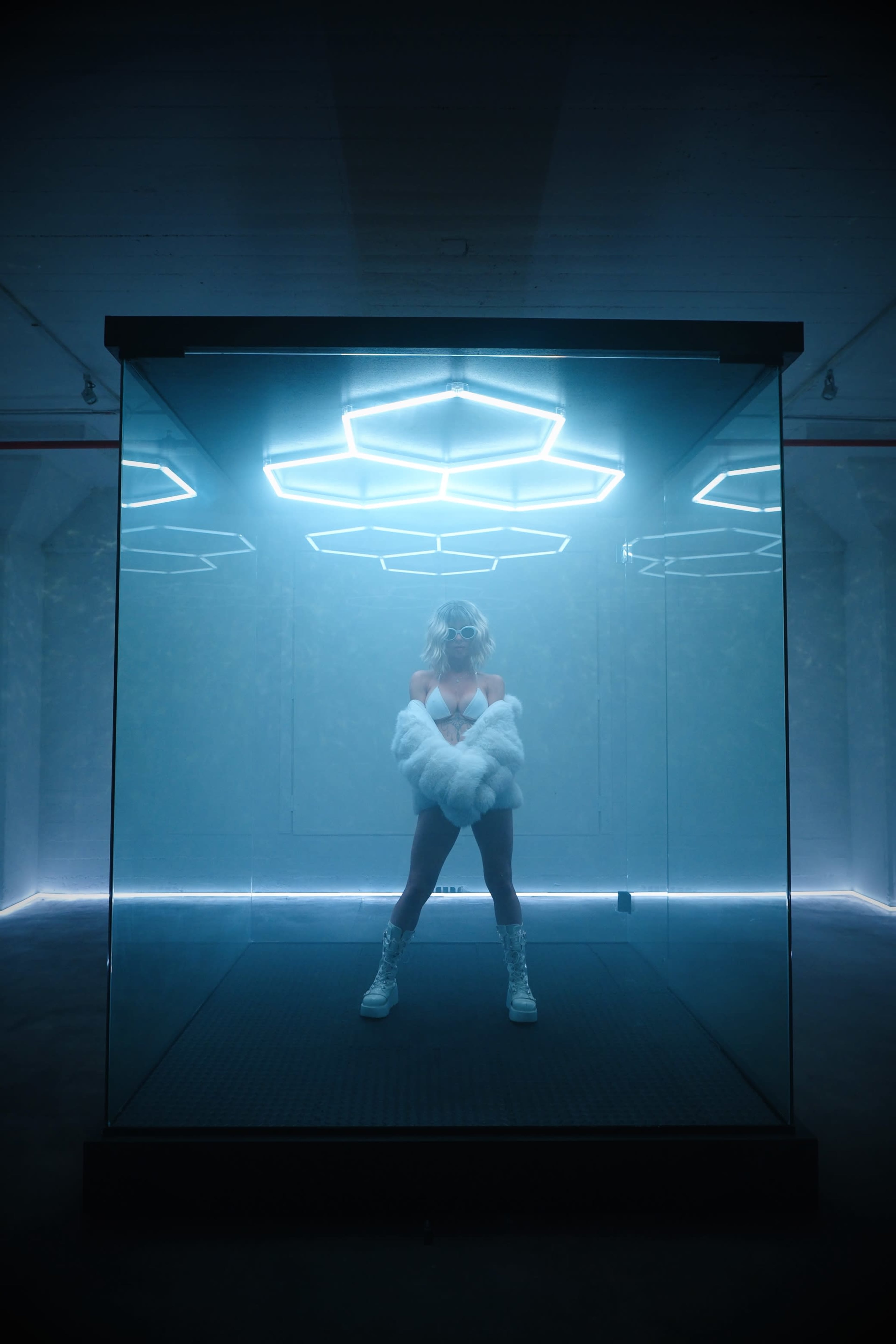 A person stands inside a glass enclosure illuminated by blue LED lights in a dimly lit room.