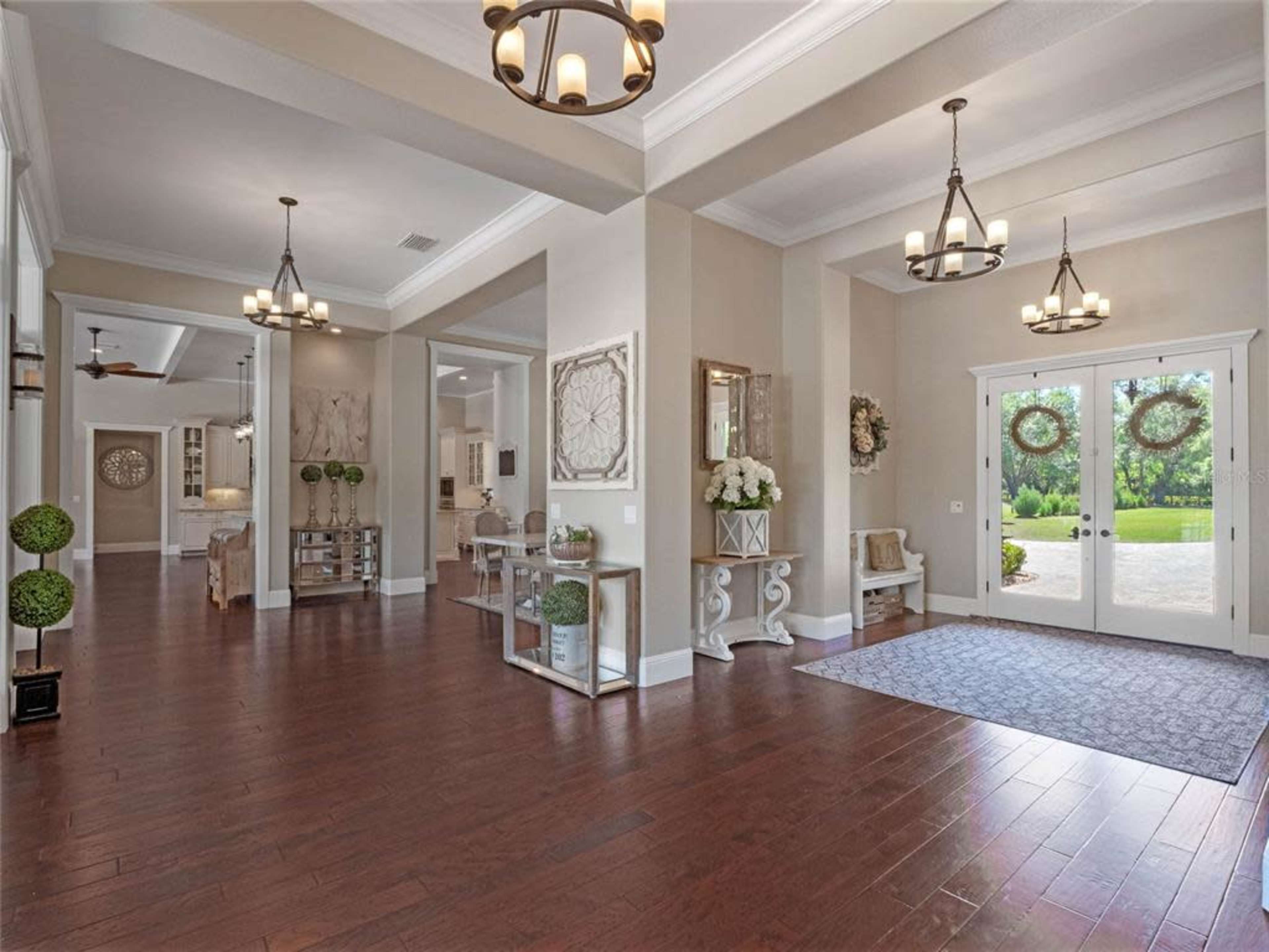 The image shows a spacious entryway with hardwood flooring, decorative chandeliers, and large double doors leading to a green outdoor area.