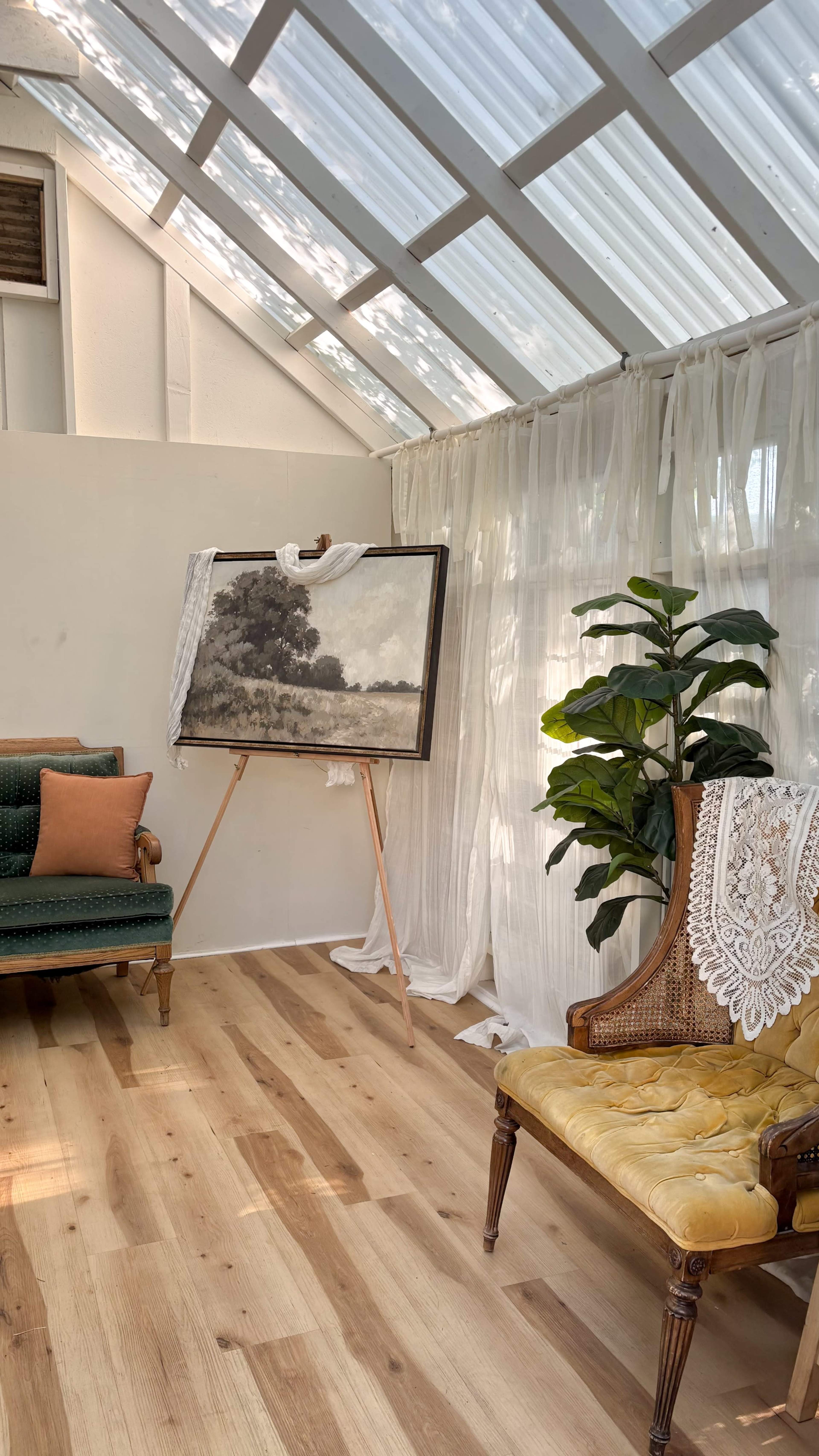 The image shows a sunlit room featuring a green upholstered sofa, a yellow chair with lace detailing, a large artwork on an easel, and a potted plant near a sheer curtain backdrop.
