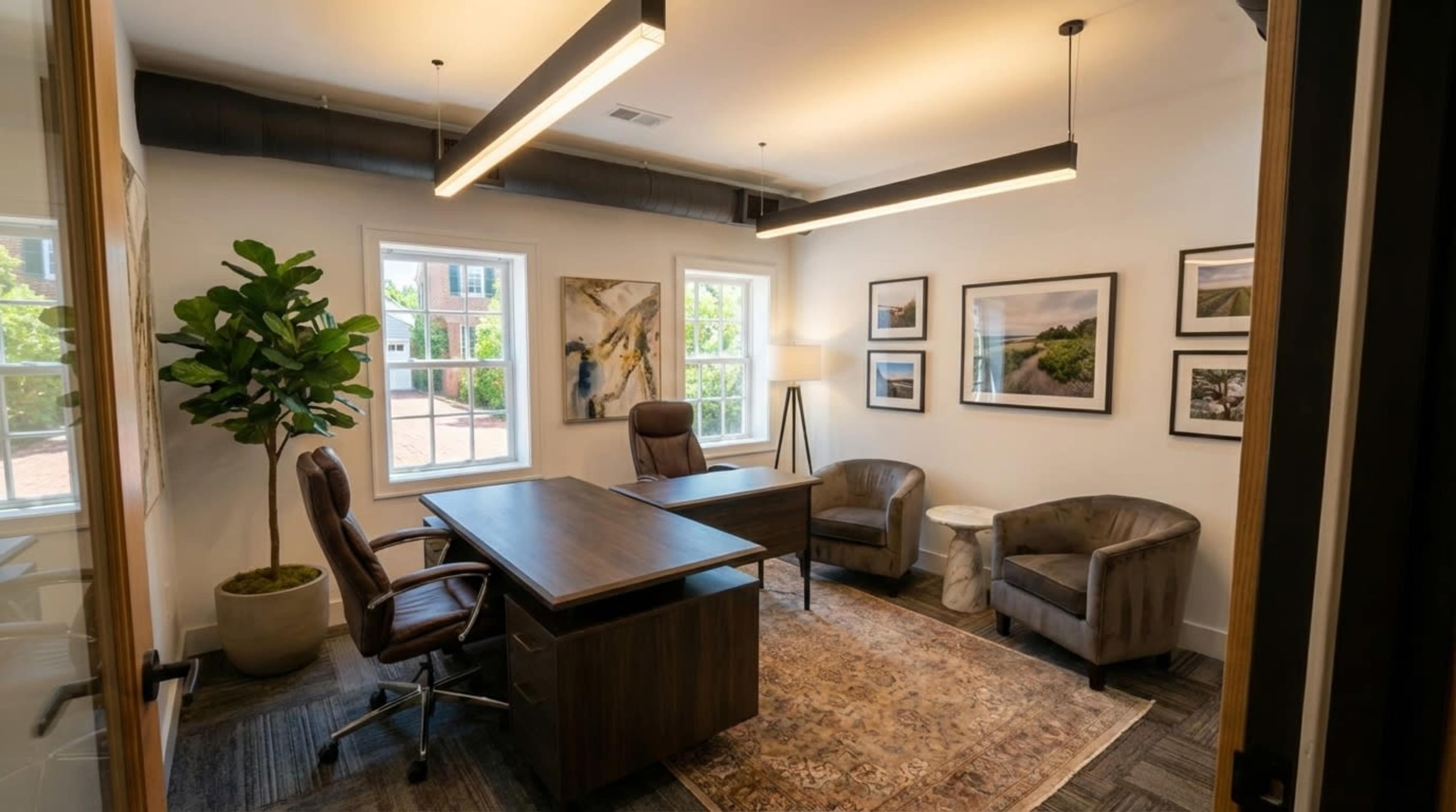The image shows a small office space with two armchairs, a desk with chairs, artwork on the walls, and a potted plant.