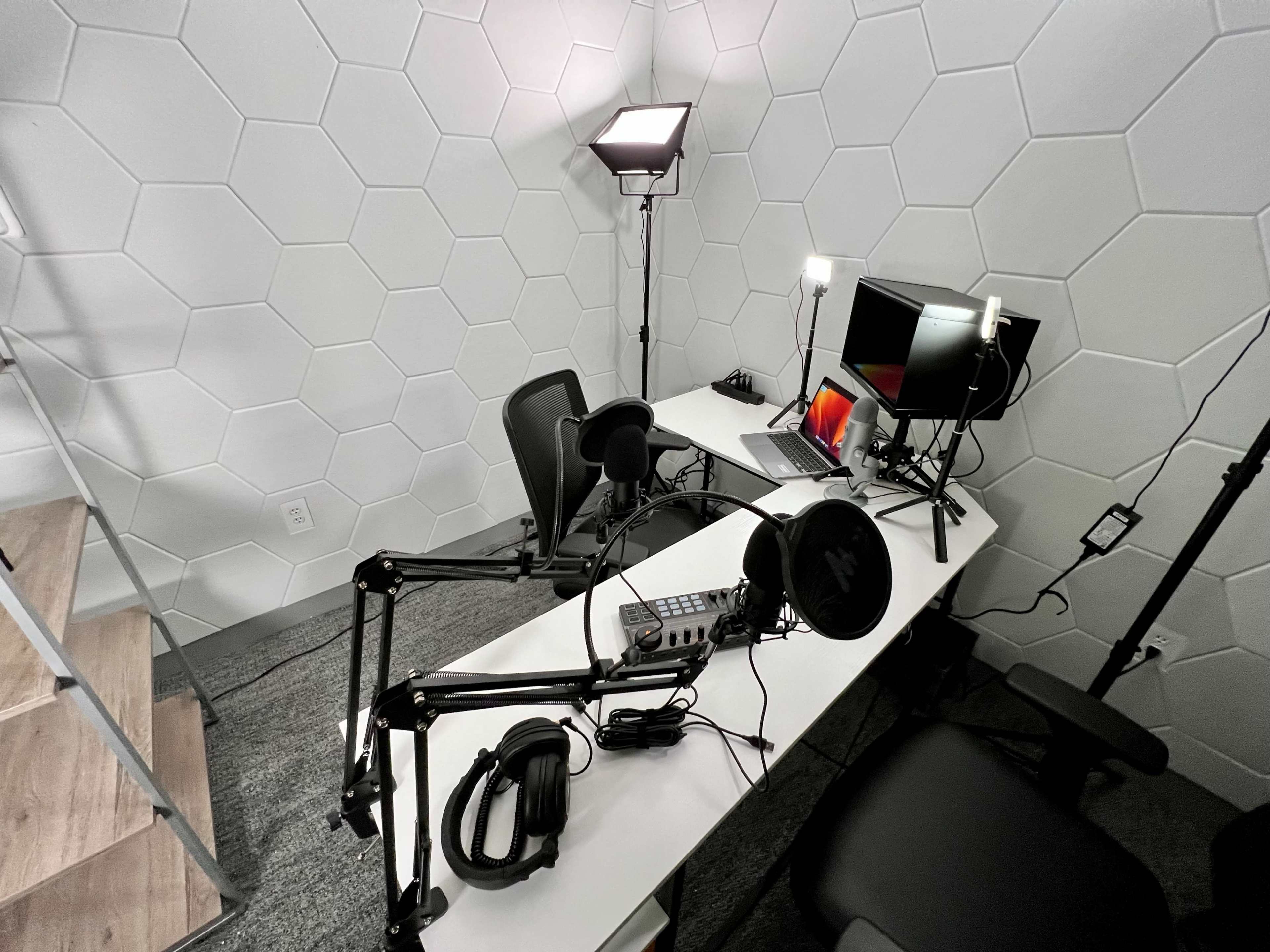 The image shows a podcasting setup featuring a desk with a computer, monitors, microphones on adjustable arms, and studio lighting in a hexagon-patterned room.