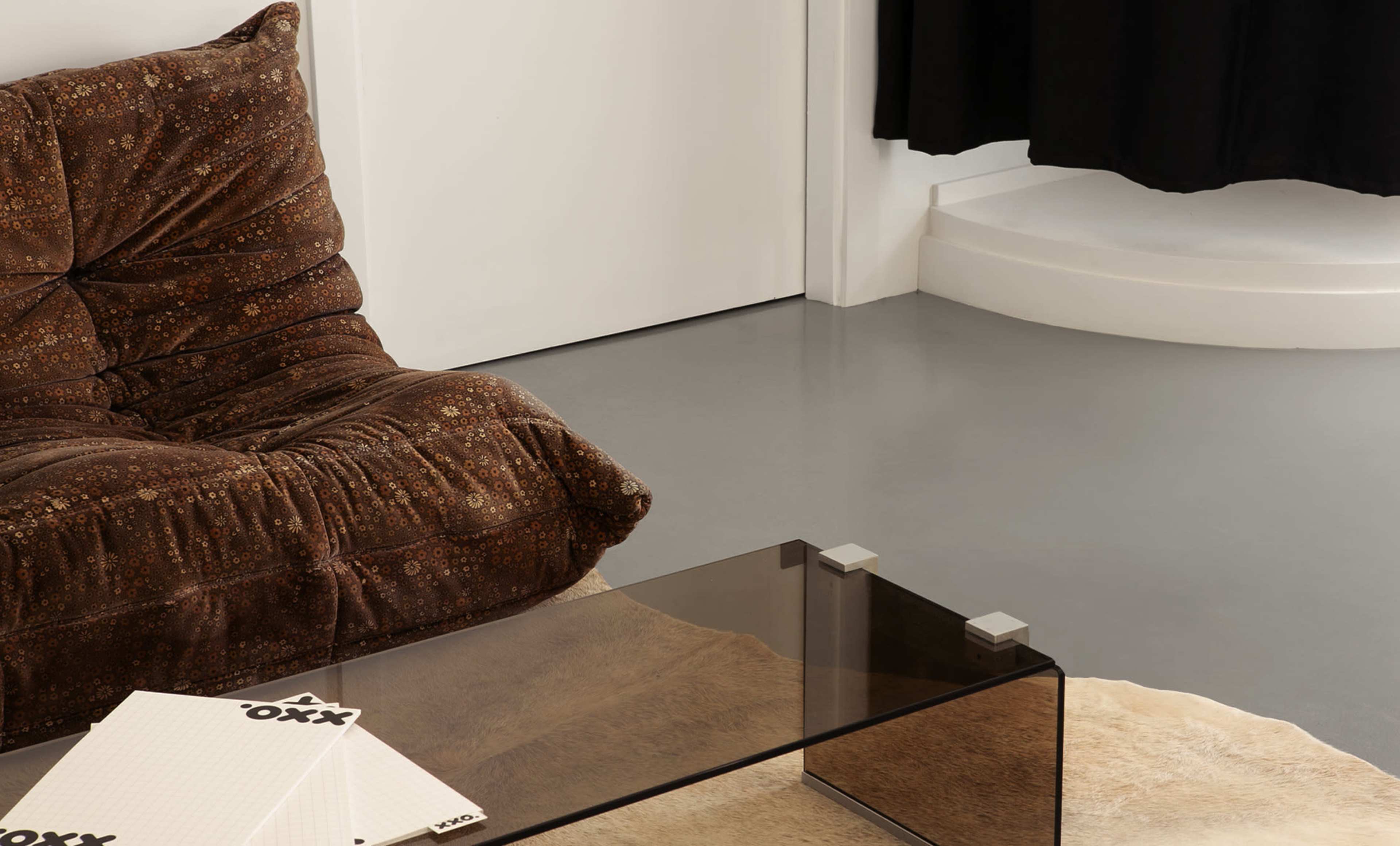 The image shows a cozy, brown upholstered chair next to a sleek glass coffee table on a polished floor.
