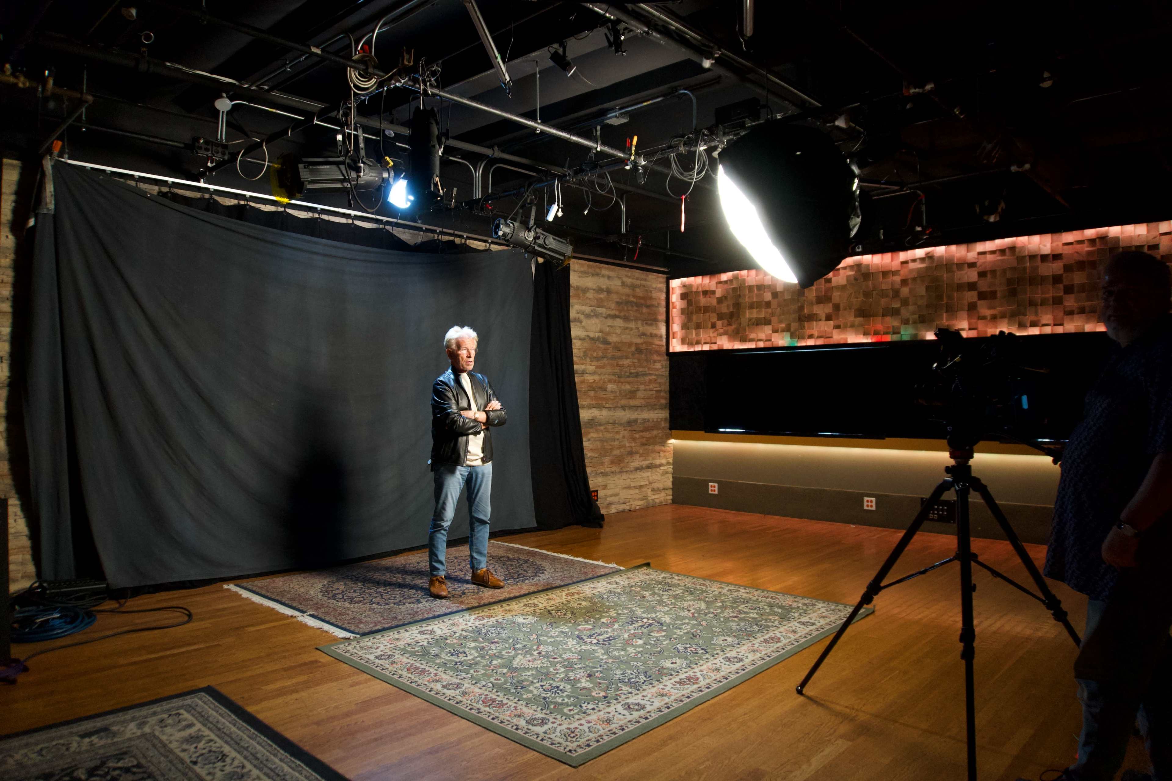 A man stands on a rug in a dimly lit studio with a black backdrop and a camera on a tripod facing him.