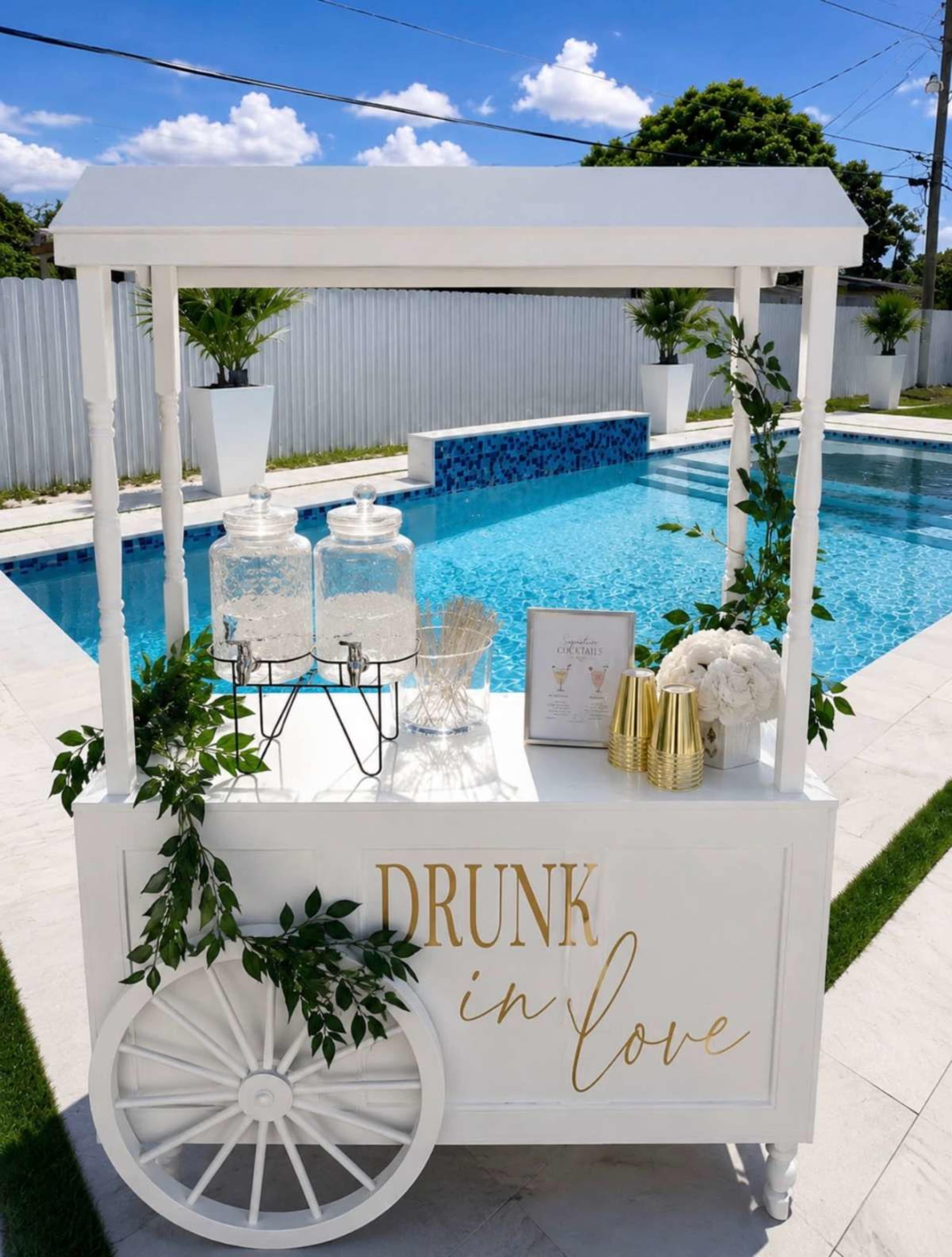 A decorative cart with the phrase "DRUNK in LOVE" is set up beside a swimming pool, featuring two glass containers, floral arrangements, and gold-colored accents.