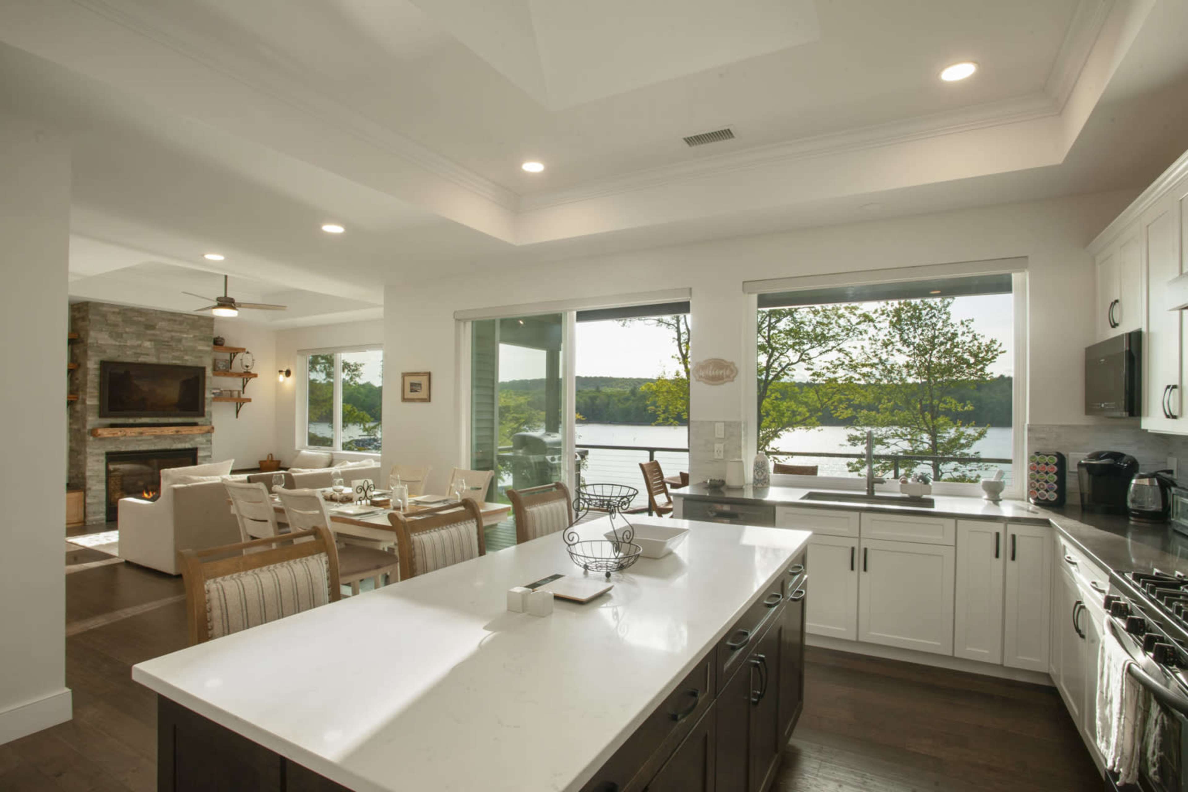 A bright kitchen features an island and opens to a living area with large windows overlooking a scenic view of water and trees.